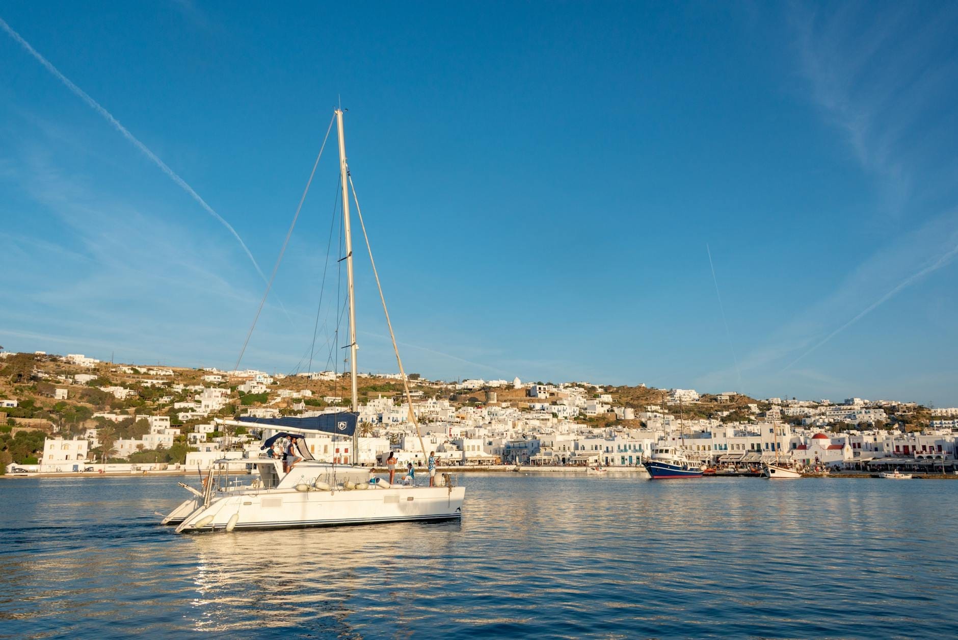 Velero en aguas tranquilas con la ciudad de Mykonos y edificios blancos tradicionales al fondo durante el día.