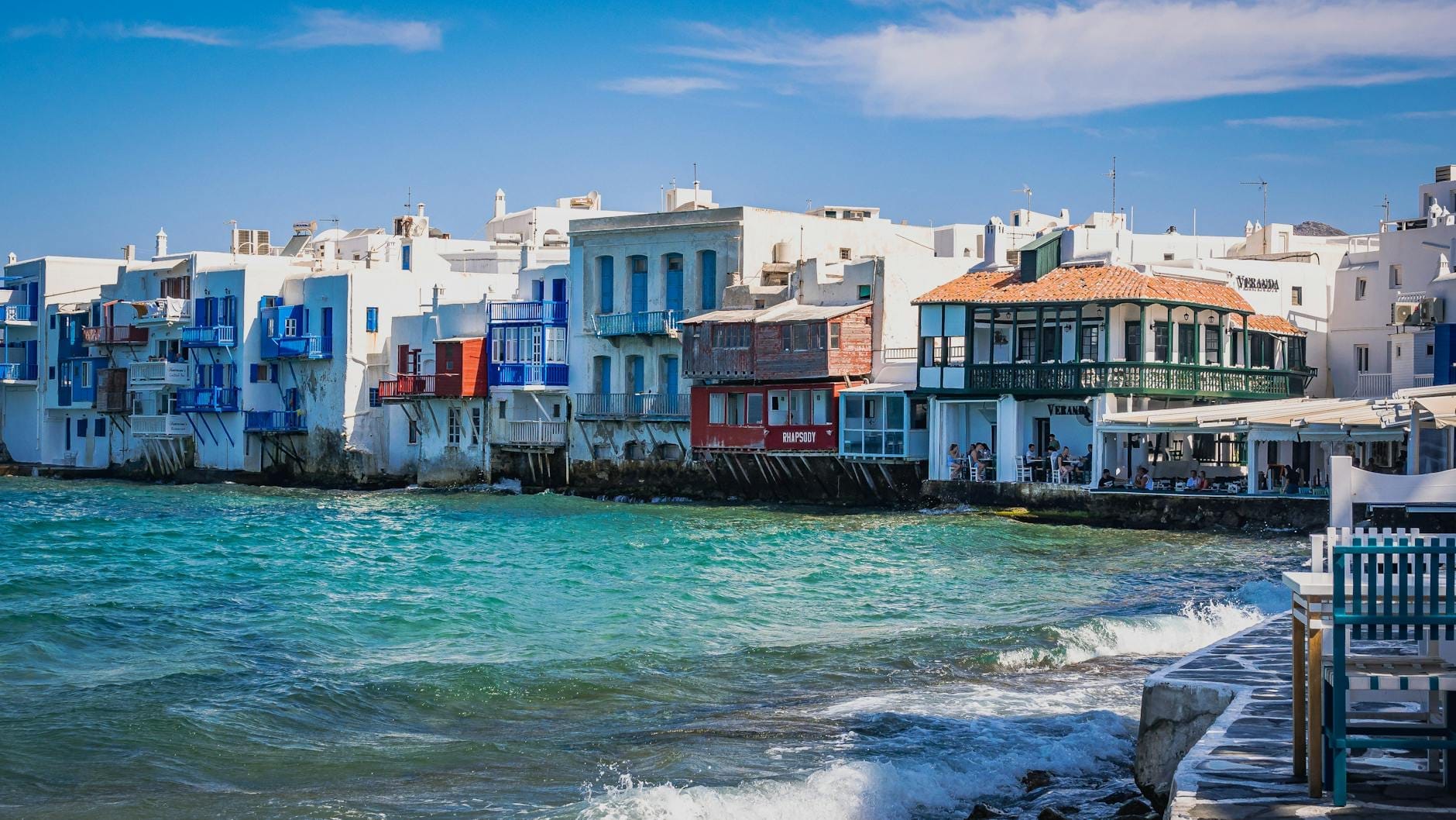 Animados bares y restaurantes frente al mar en Little Venice de Mykonos, con olas rompiendo y gente relajándose en las mesas al aire libre.