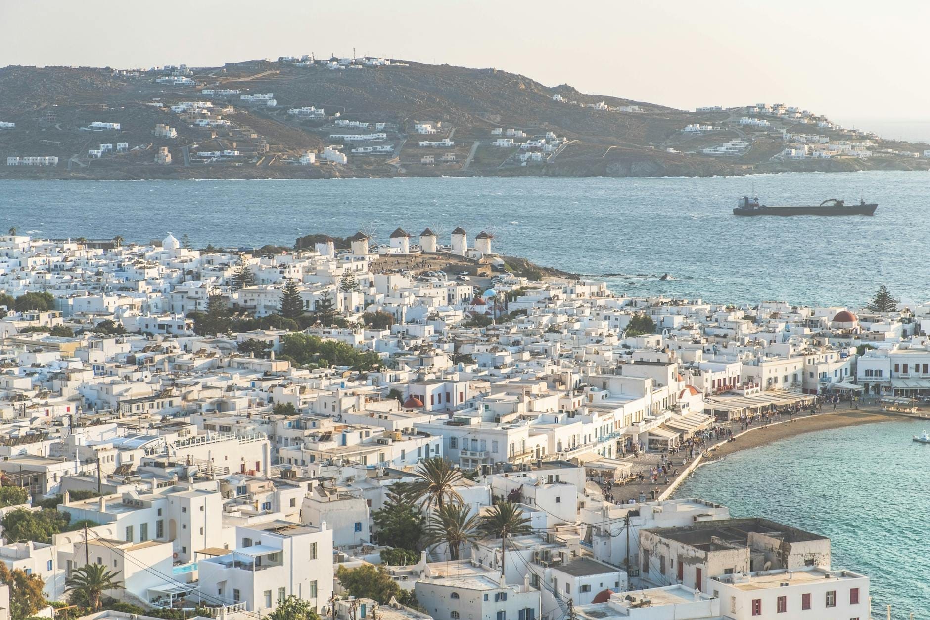 Vista aérea amplia de la ciudad de Mykonos con edificios encalados, costa, puerto y los icónicos molinos de viento, con colinas y mar al fondo.