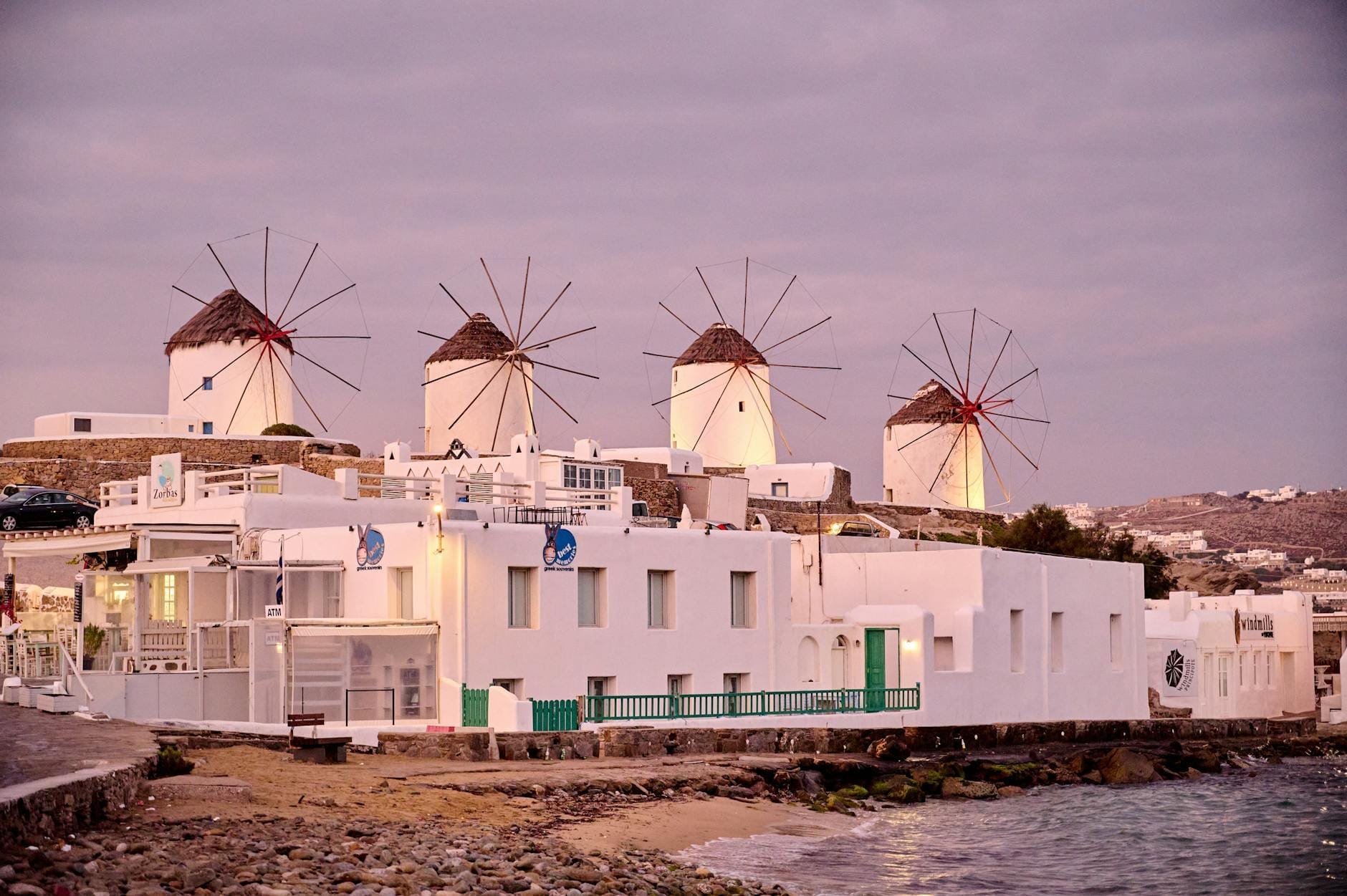 Cuatro icónicos molinos de viento encalados con techos de paja se alzan sobre una colina sobre el pueblo de Mykonos, con vistas a edificios blancos y el mar Egeo al atardecer.