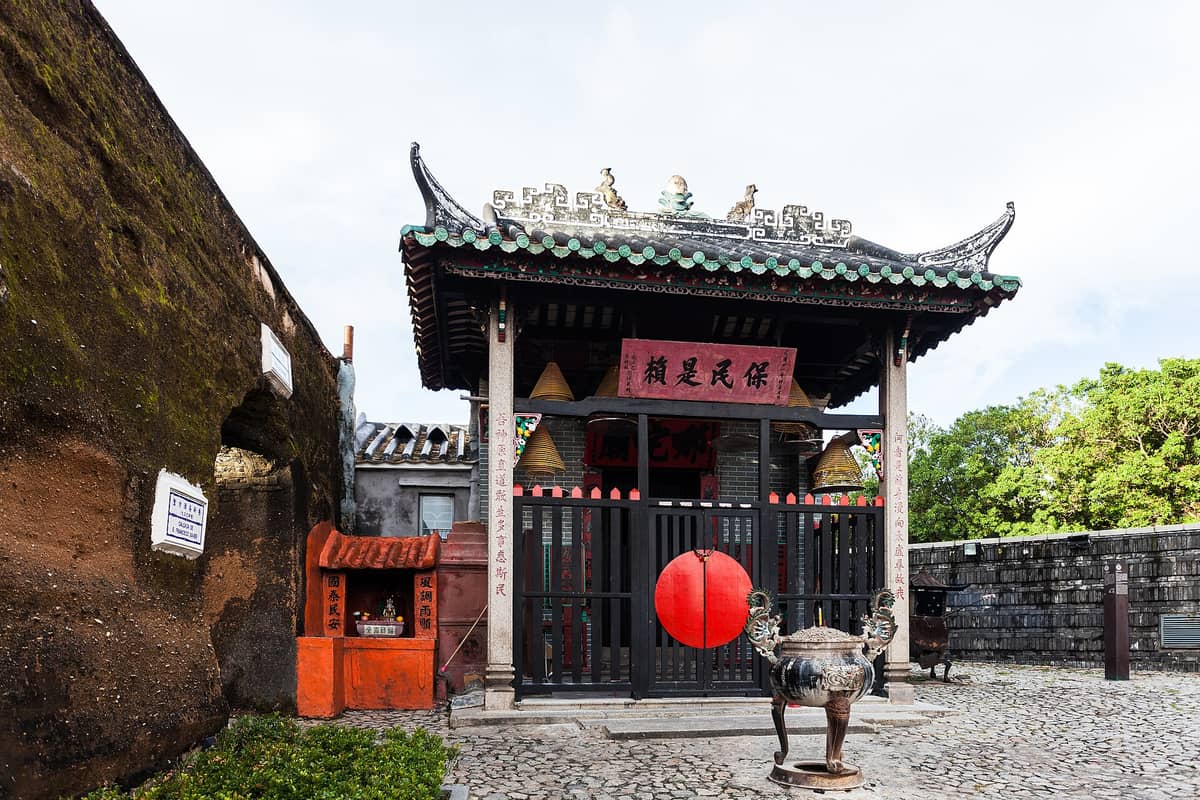 Der Na-Tcha-Tempel in Macau – ein kleiner traditioneller chinesischer Tempel mit grünem Ziegeldach und hängenden Räucherspiralen, direkt an die alte Steinmauer des St.-Pauls-Kollegs geschmiegt.