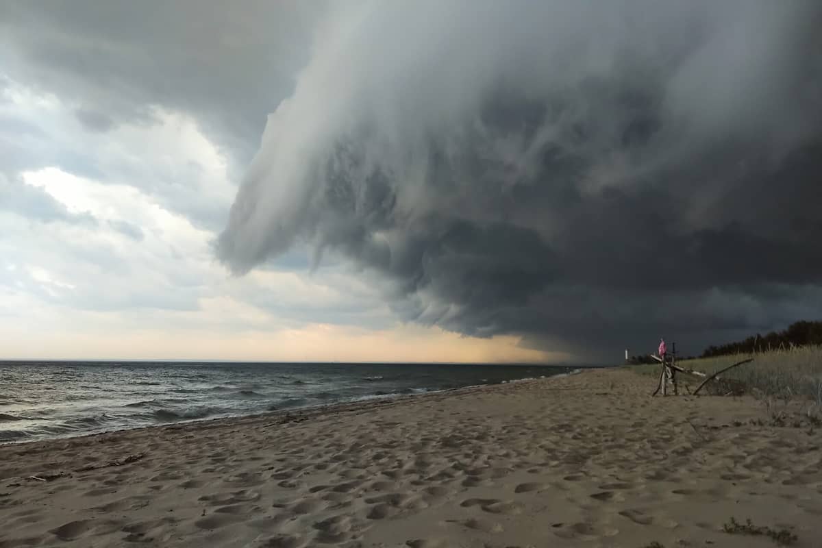 Sandstrand auf Naissaar mit dunklen Gewitterwolken, Wellen am Ufer und einem Leuchtturm in der Ferne am Rand des Kiefernwaldes.
