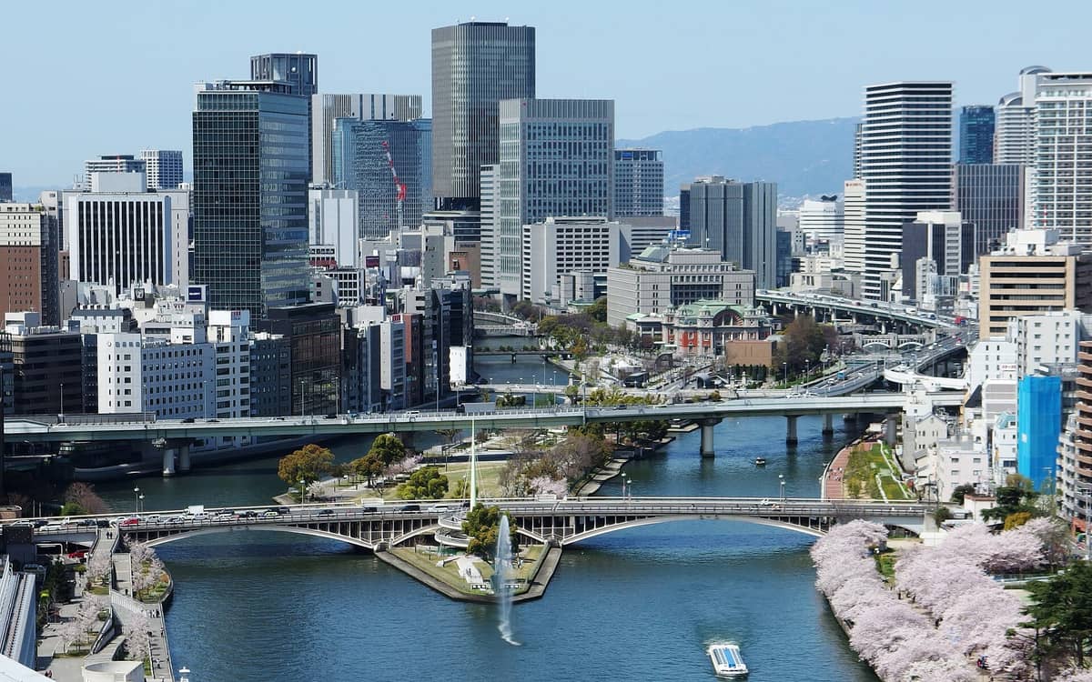 A panoramic aerial view of Nakanoshima island in central Osaka with bridges, rivers, cherry blossoms, and the city skyline featuring Meiji-era architecture and modern buildings.
