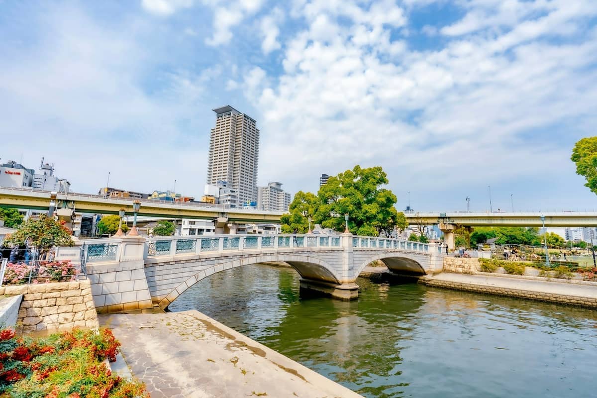 Stone pedestrian bridge crossing the river at Nakanoshima Park, with city buildings, trees, and bright blue sky in Osaka, Japan.