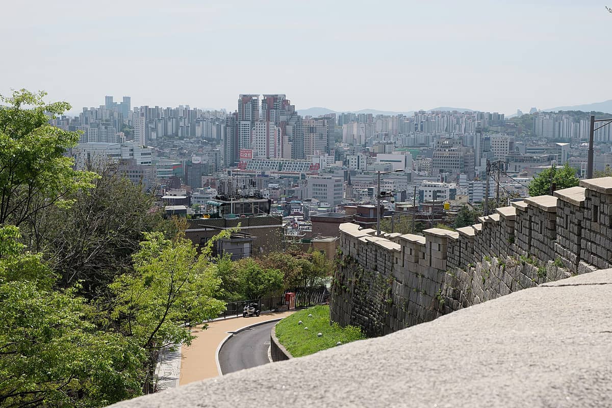 Muralha de pedra do Parque Naksan descendo a colina em curva, com os prédios de Seul ao fundo num dia de céu limpo.