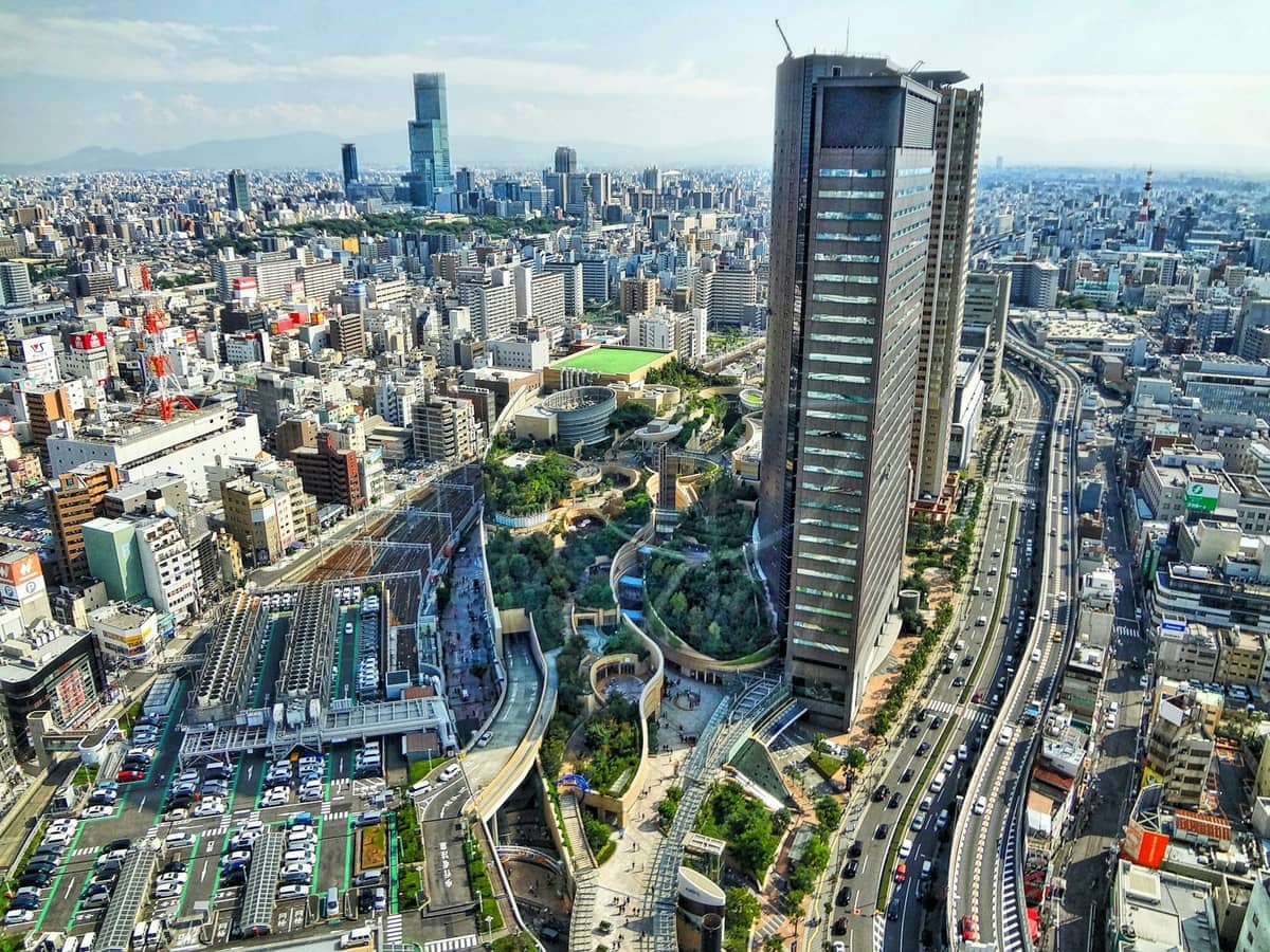 Panoramic aerial view of Namba, Osaka, featuring high-rise buildings, busy roads, railway tracks, and urban green spaces under a clear sky.