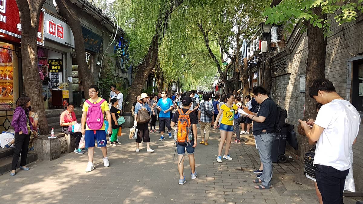 Belebte Fußgängergasse in Nanluoguxiang, Peking, mit traditionellen grauen Backsteingebäuden, Weidenbäumen, Ladenschildern und Menschen, die das historische Viertel erkunden.