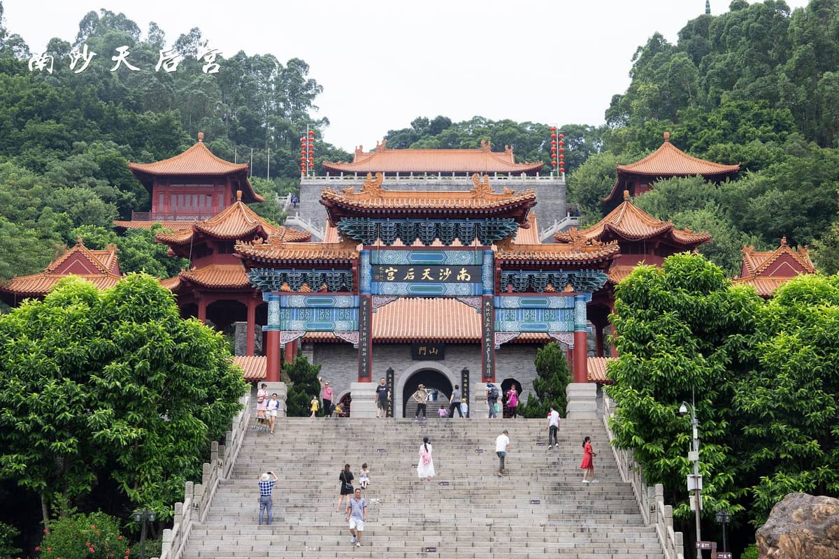 Front view of Nansha Tianhou Palace showing grand staircase, traditional architecture with orange tiled roofs, lush green trees, and people ascending the temple steps.