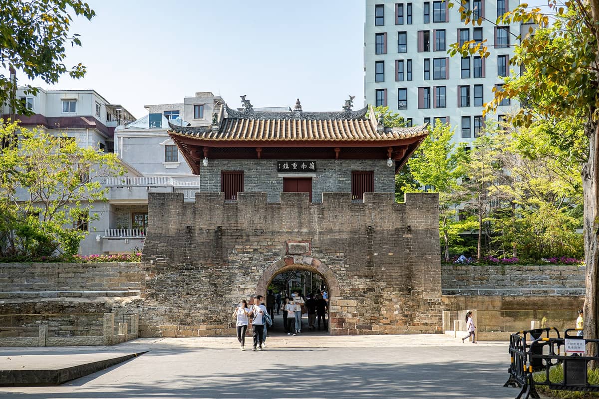 Vista frontal do portão principal e da muralha de pedra da Cidade Antiga de Nantou, em Shenzhen, com pessoas passando sob o arco e prédios modernos ao fundo.