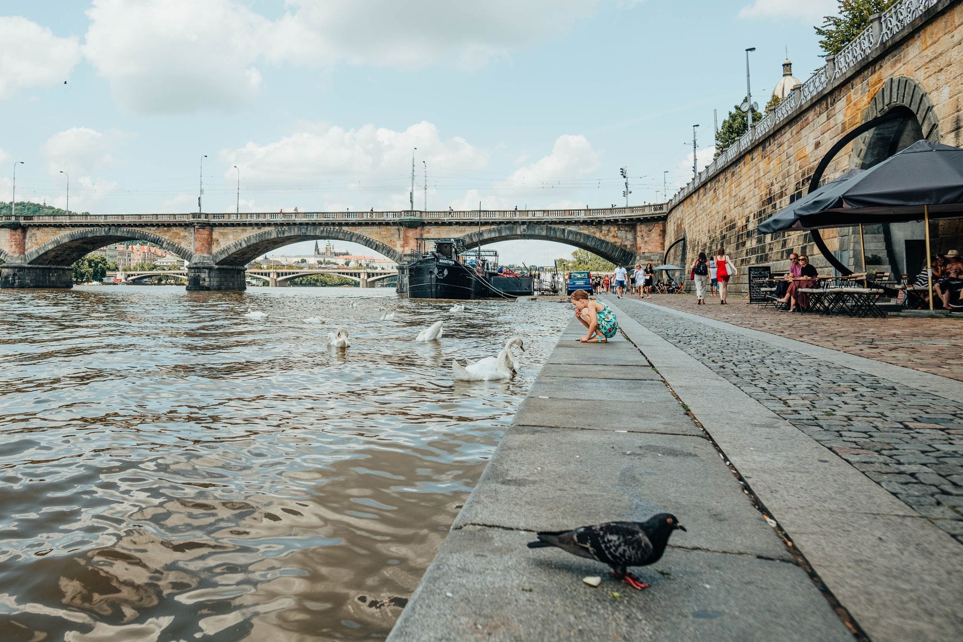 Blick entlang der Náplavka-Uferpromenade mit Kopfsteinpflaster, Menschen am Wasser, Schwänen, festgemachten Booten und einer historischen Steinbrücke über die Moldau in Prag.
