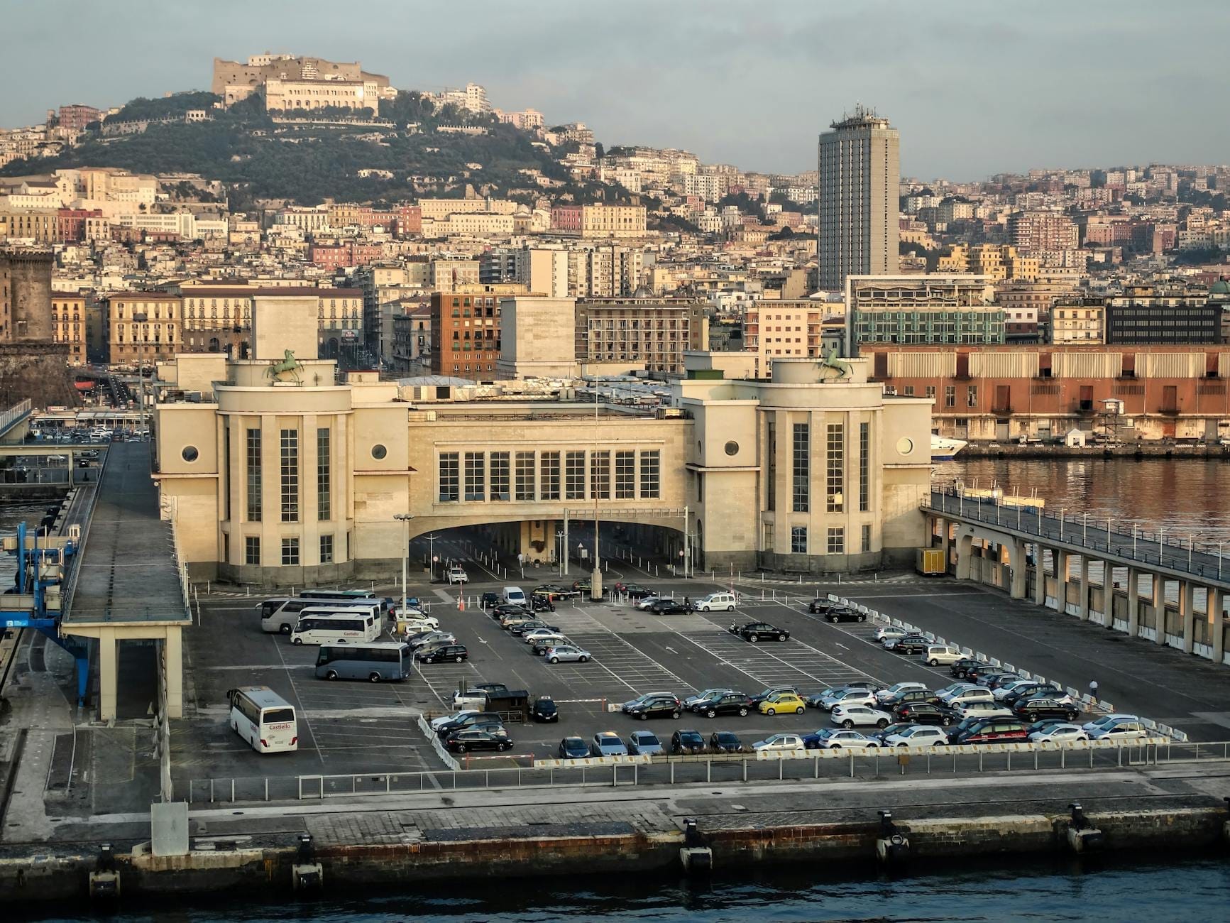 Parking devant le terminal du port de Naples avec des voitures, des camionnettes et des bus visibles, sur fond de paysage urbain et de collines de Naples en plein jour.
