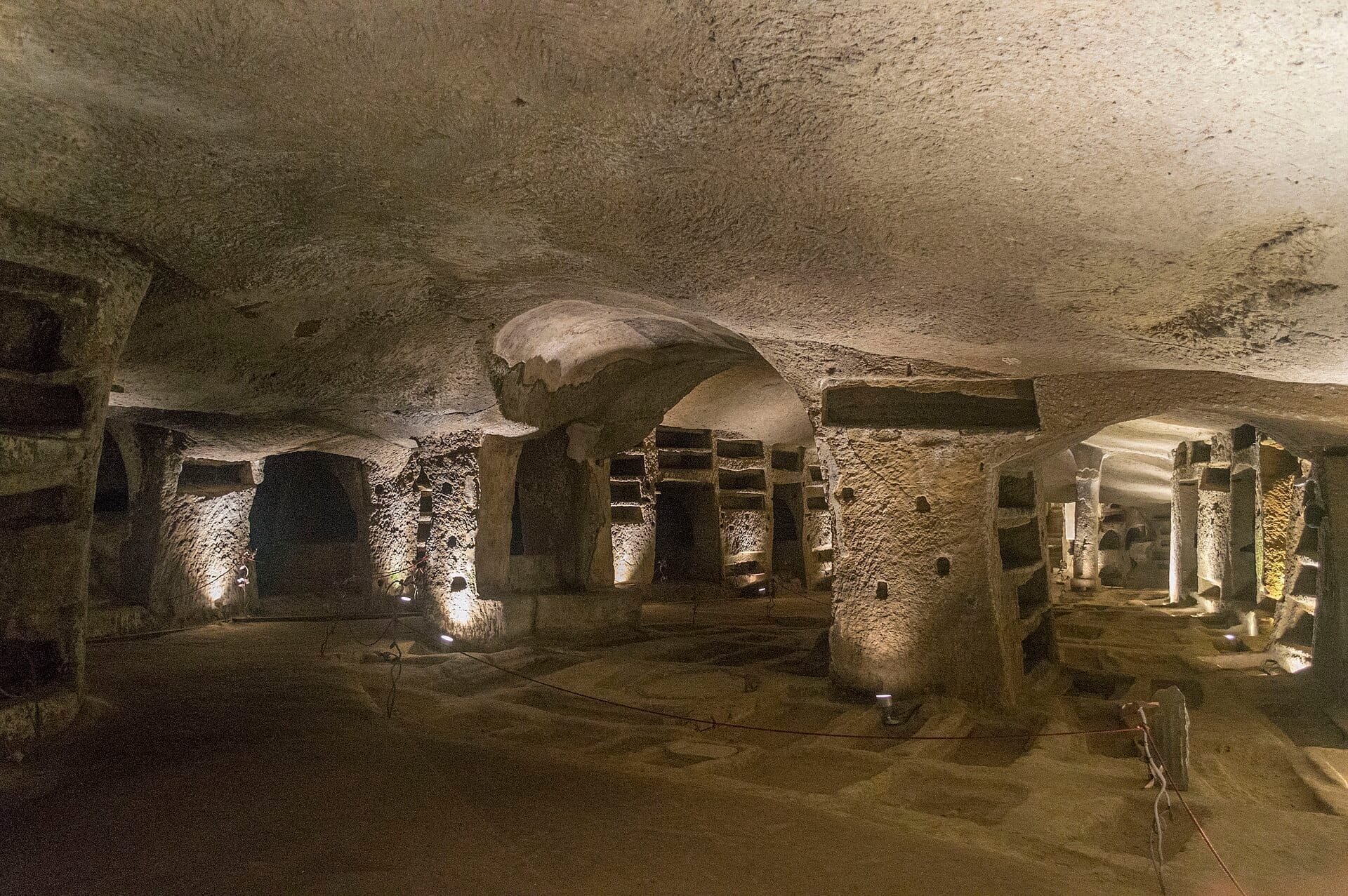 Vista ampia delle Catacombe di San Gennaro illuminate, con pilastri in pietra scolpita, soffitti ad arco e nicchie sepolcrali nella città sacra sotterranea di Napoli.