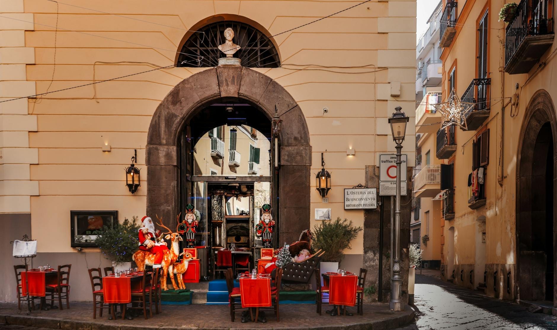 Pequena praça em Nápoles com decorações natalinas festivas, toalhas de mesa vermelhas e figuras de Papai Noel em frente à entrada de um restaurante.