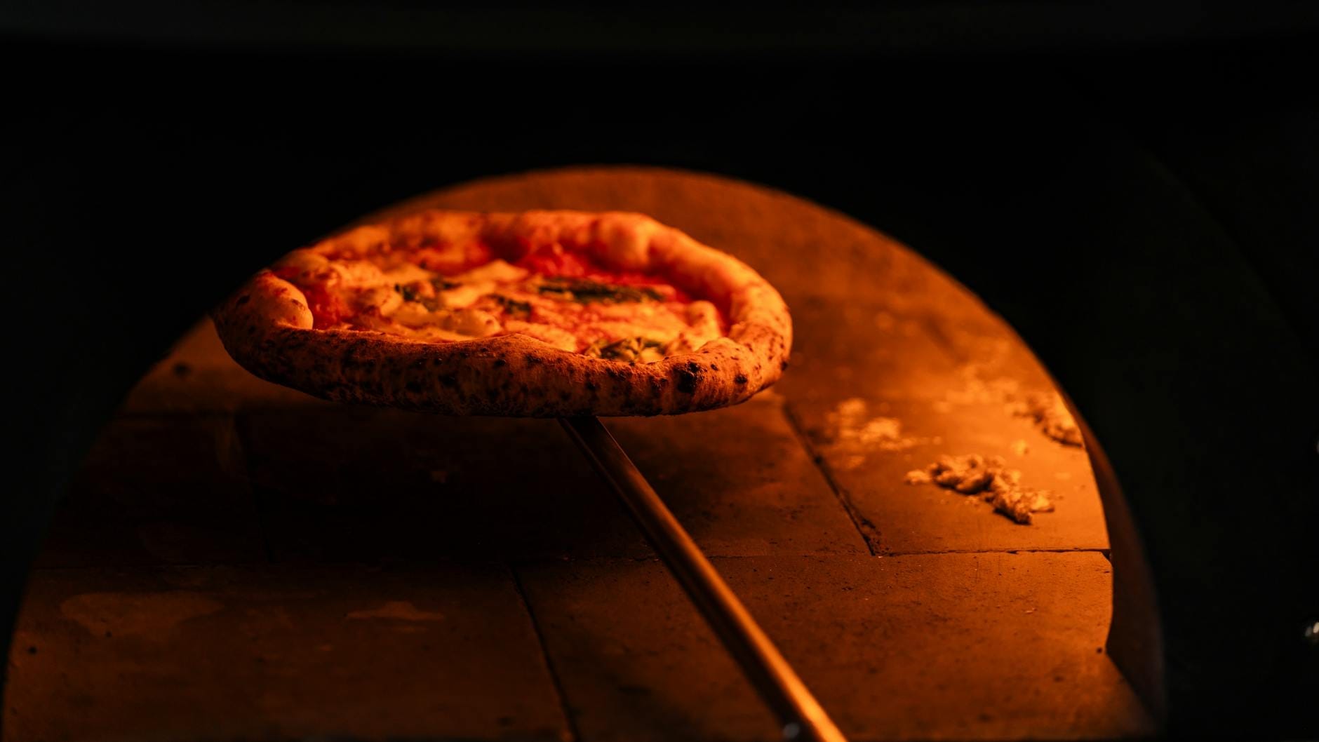 Vue rapprochée d'une pizza napolitaine en train de cuire dans un four à bois, avec des flammes visibles et une croûte légèrement brûlée.