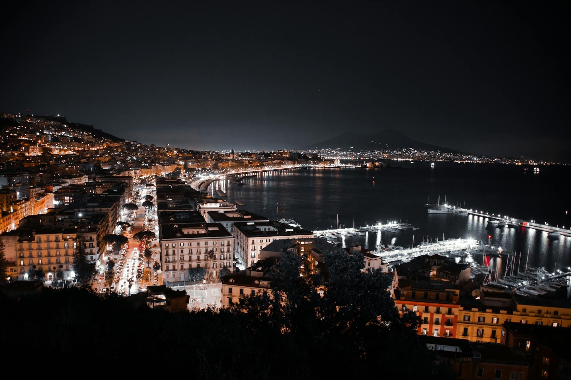 Vista aérea noturna do litoral de Nápoles com as luzes da cidade refletidas na baía e o Monte Vesúvio ao fundo.