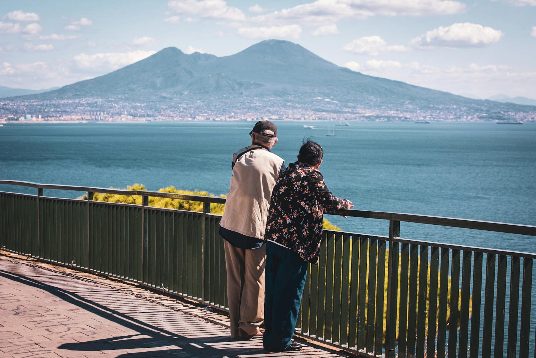 Casal mais velho apoiado numa grade, apreciando a vista do mar de Nápoles com o Monte Vesúvio ao fundo.