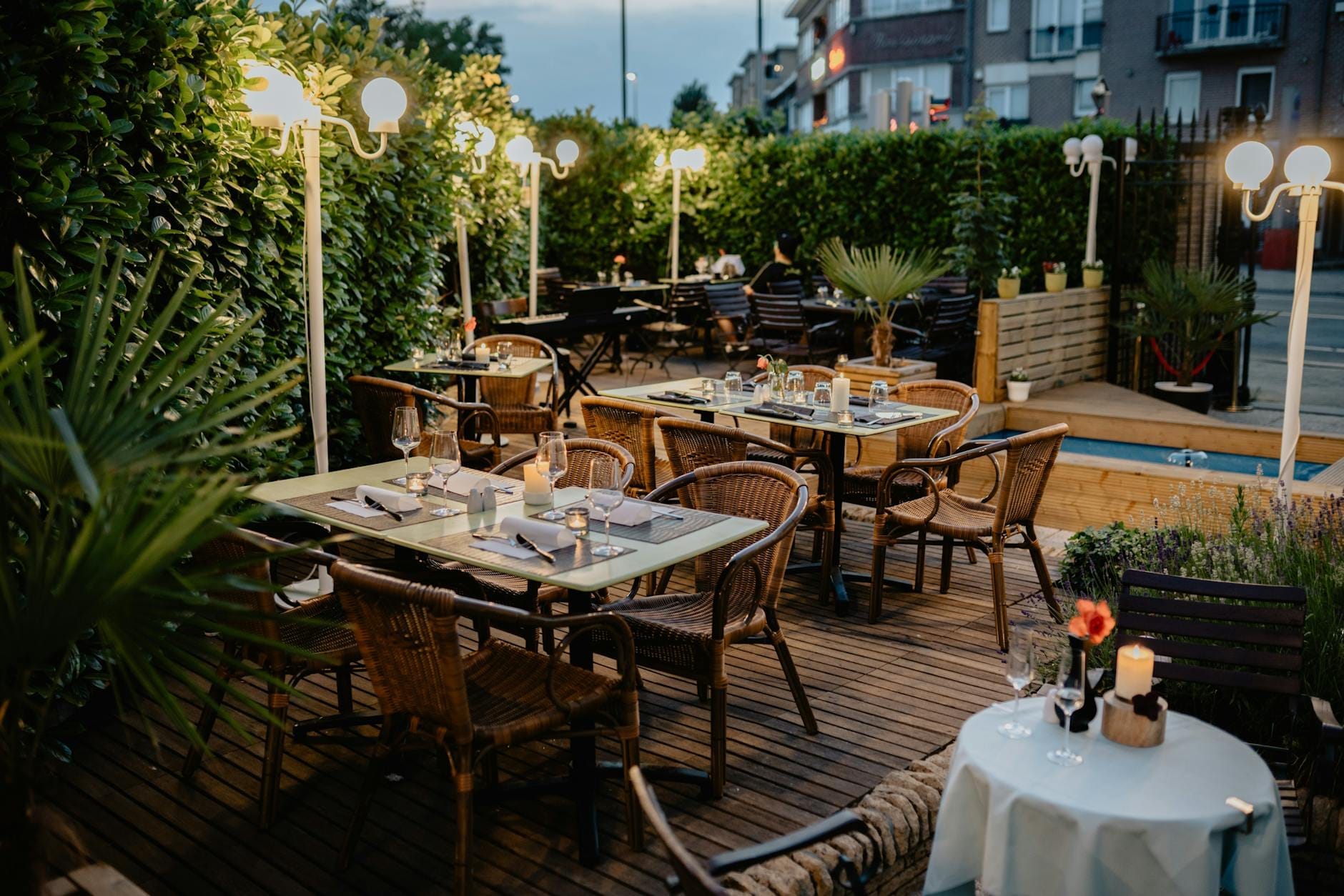 Pátio de restaurante ao ar livre com mesas arrumadas, cadeiras de vime e velas, rodeado de vegetação num ambiente urbano europeu ao entardecer.