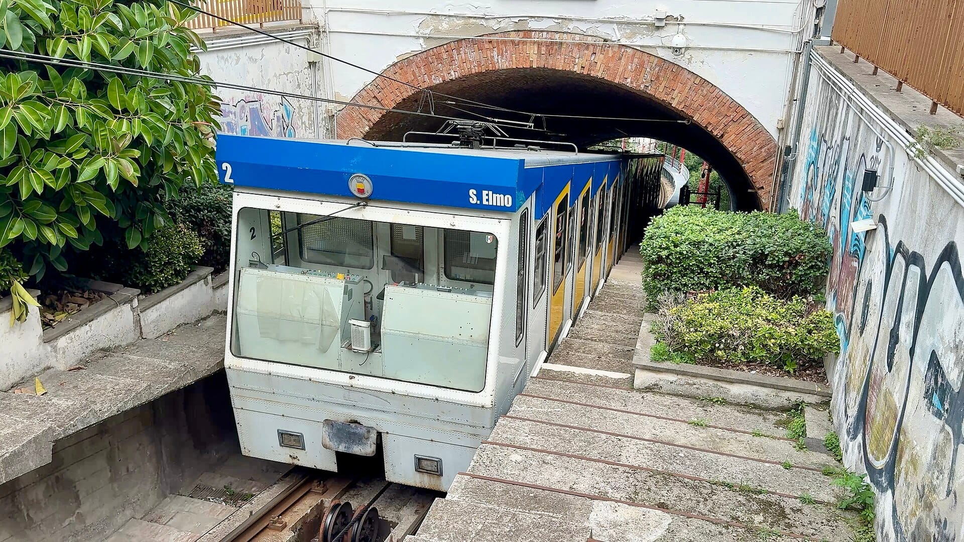 La carrozza della funicolare di Napoli S. Elmo esce da un tunnel di mattoni, fiancheggiata da cespugli verdi e muri con graffiti sotto una luce diurna nuvolosa.