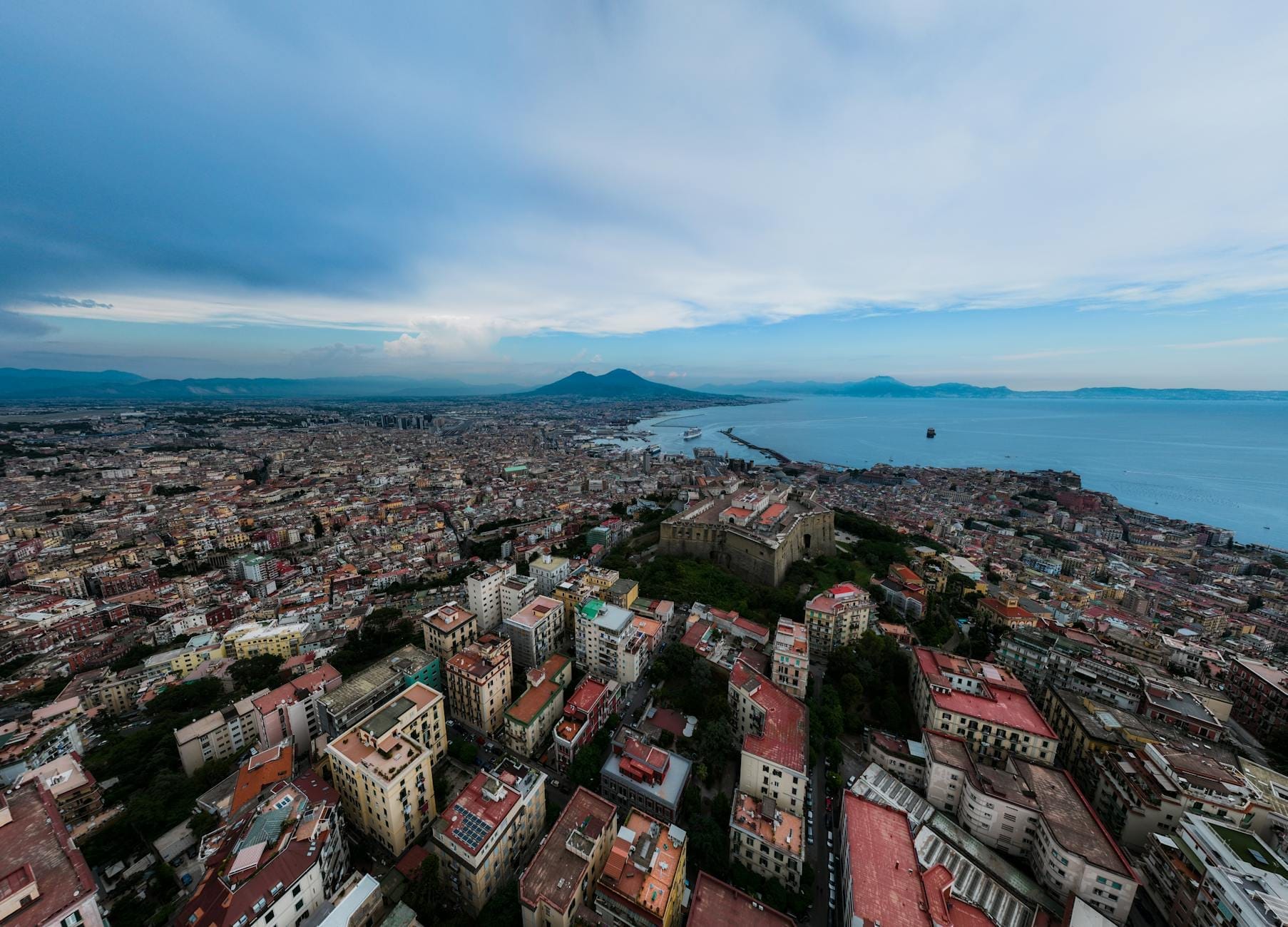 Vue aérienne de Naples avec la mer au sud, le Vésuve au loin et les bâtiments de la ville s'étendant en dessous sous un ciel bleu.
