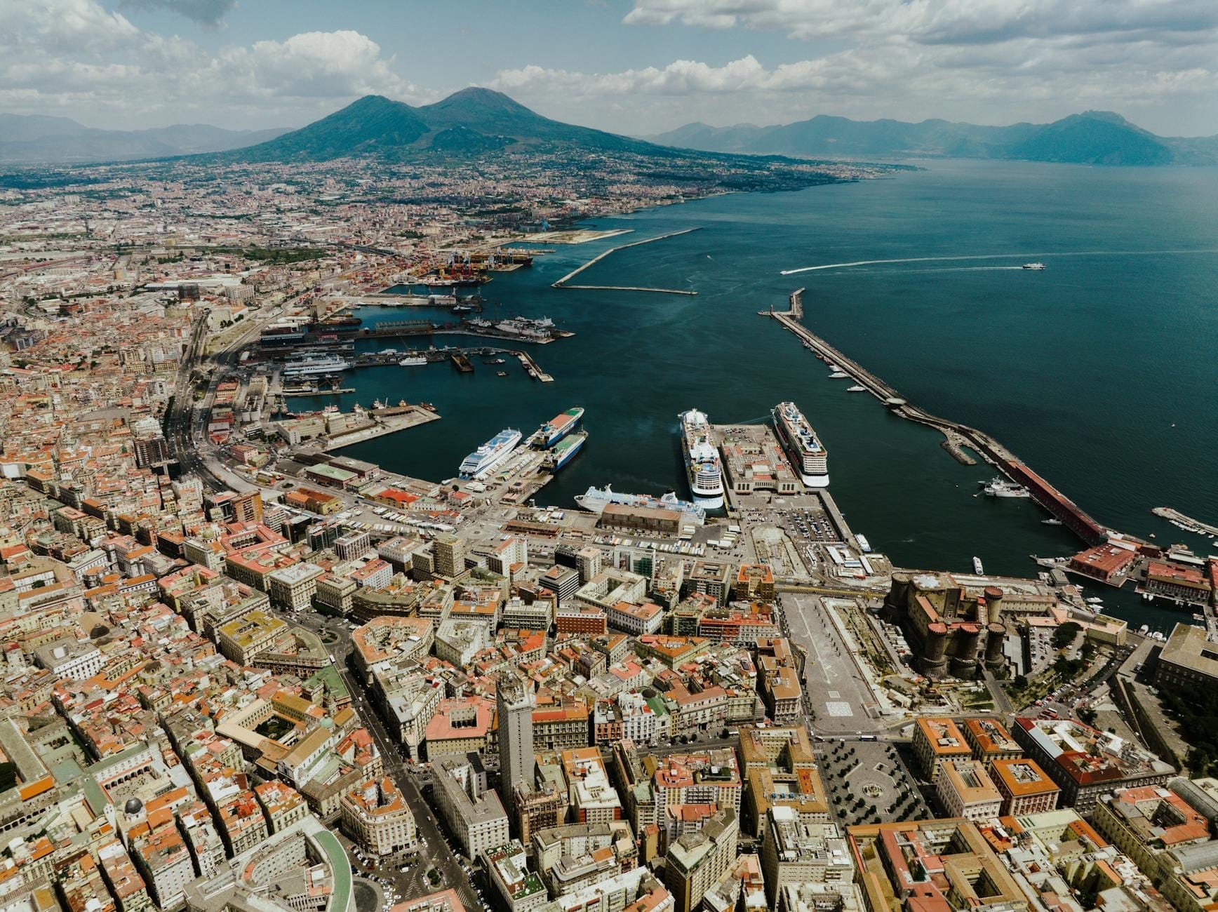 Vista aérea panorâmica de Nápoles mostrando os bairros da cidade, a área do porto e o Monte Vesúvio ao fundo em um dia de céu limpo.