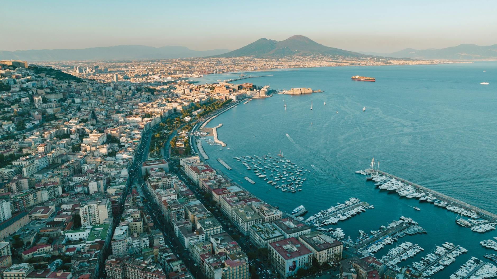 Vue aérienne du front de mer de Naples avec le Lungomare, le port de plaisance et le Vésuve en arrière-plan par une journée claire.