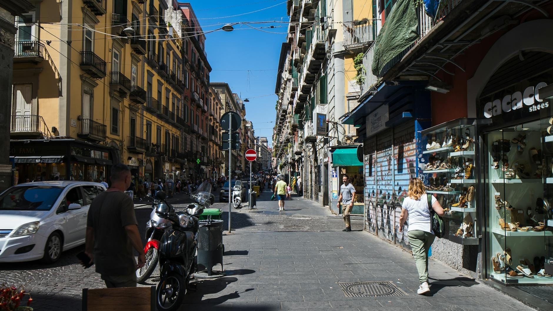 Une rue animée dans le centre historique de Naples avec des boutiques, des passants, des scooters et de vieux bâtiments de chaque côté de la rue.