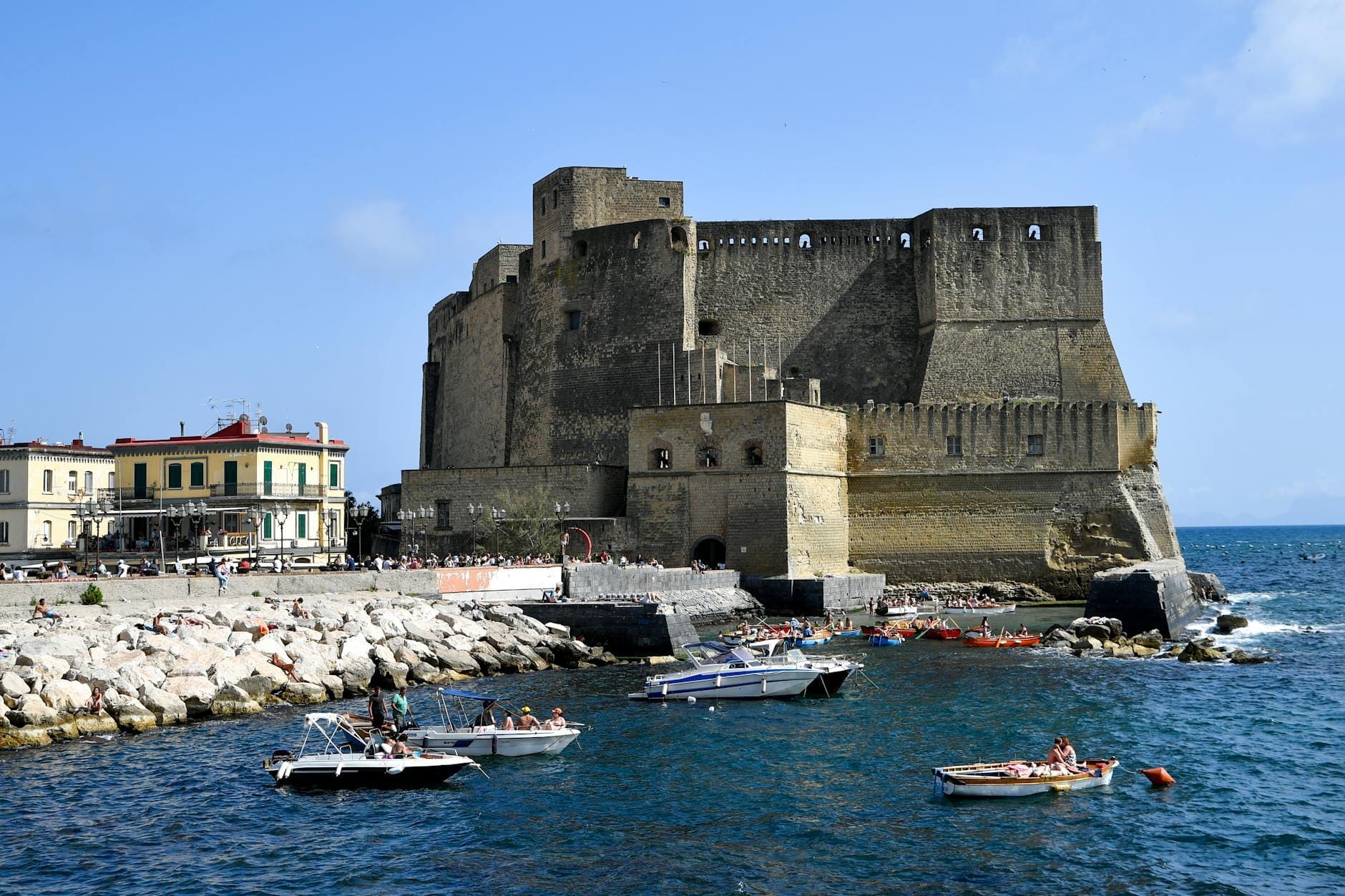 Le Castel dell'Ovo sur le front de mer de Naples avec de petits bateaux dans une eau bleue et des personnes profitant du rivage rocheux.