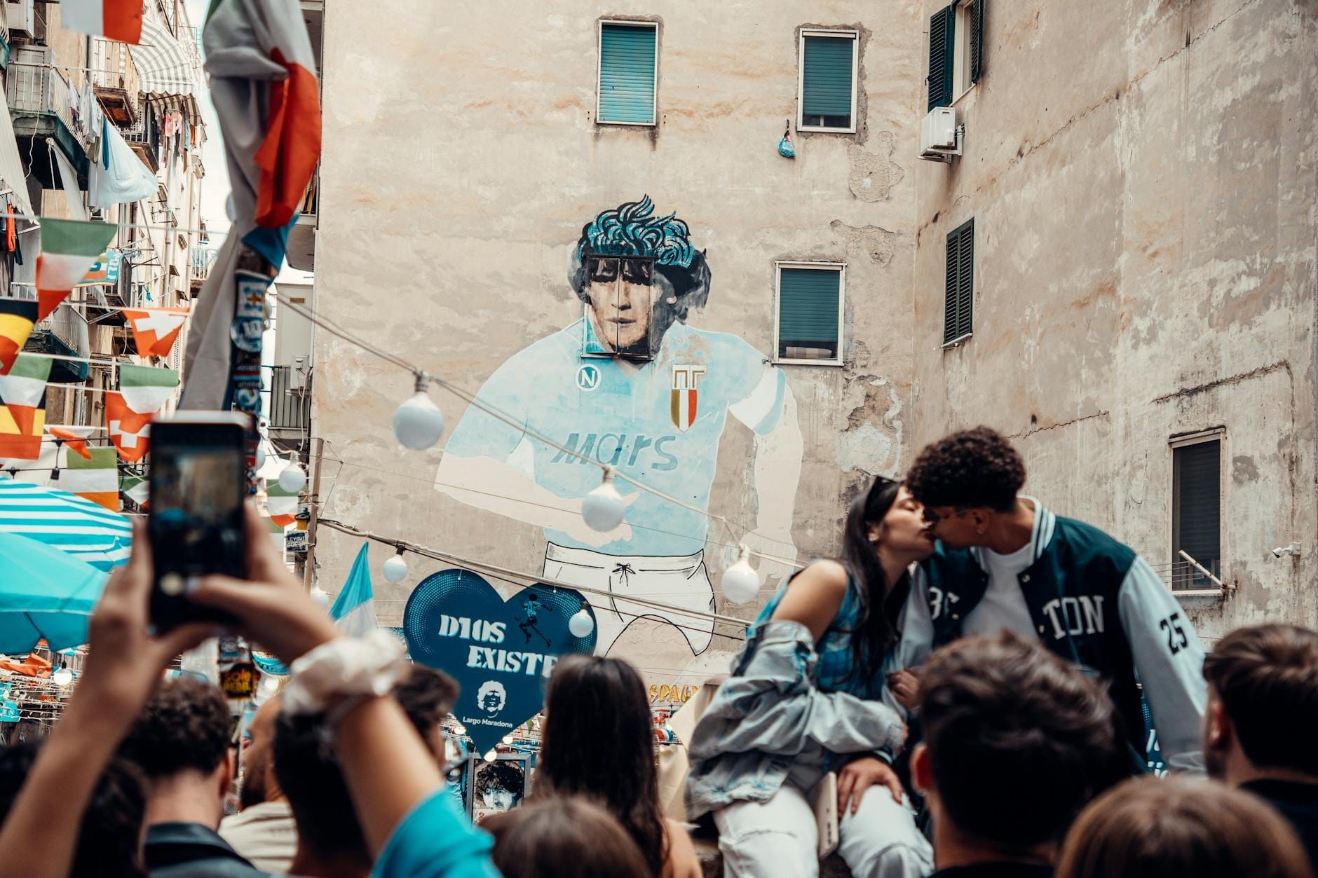 Una vivace scena di strada a Napoli con persone riunite attorno a un grande murales di Maradona su un palazzo, circondato da bandiere e decorazioni colorate.