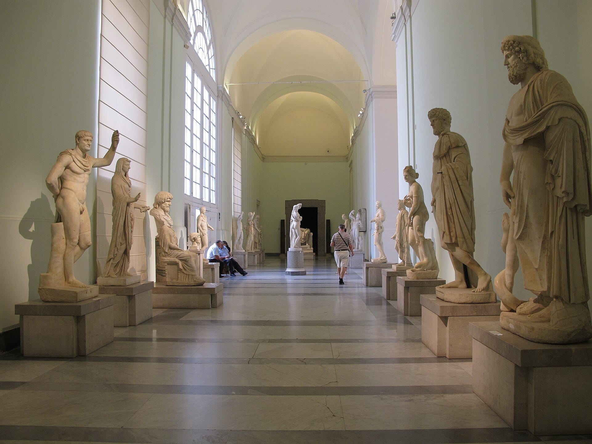 Galleria di statue in marmo al Museo Archeologico Nazionale di Napoli, con visitatori che passeggiano e luce naturale che filtra attraverso grandi finestre.