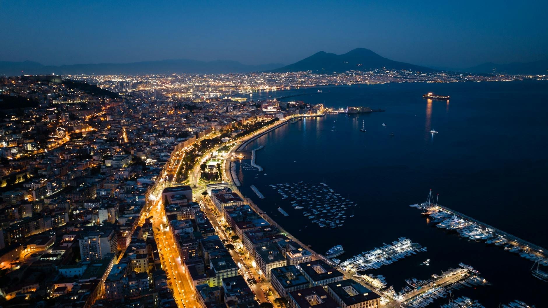 Vista aérea do litoral de Nápoles à noite com luzes da cidade, marina e o Monte Vesúvio visível ao fundo.