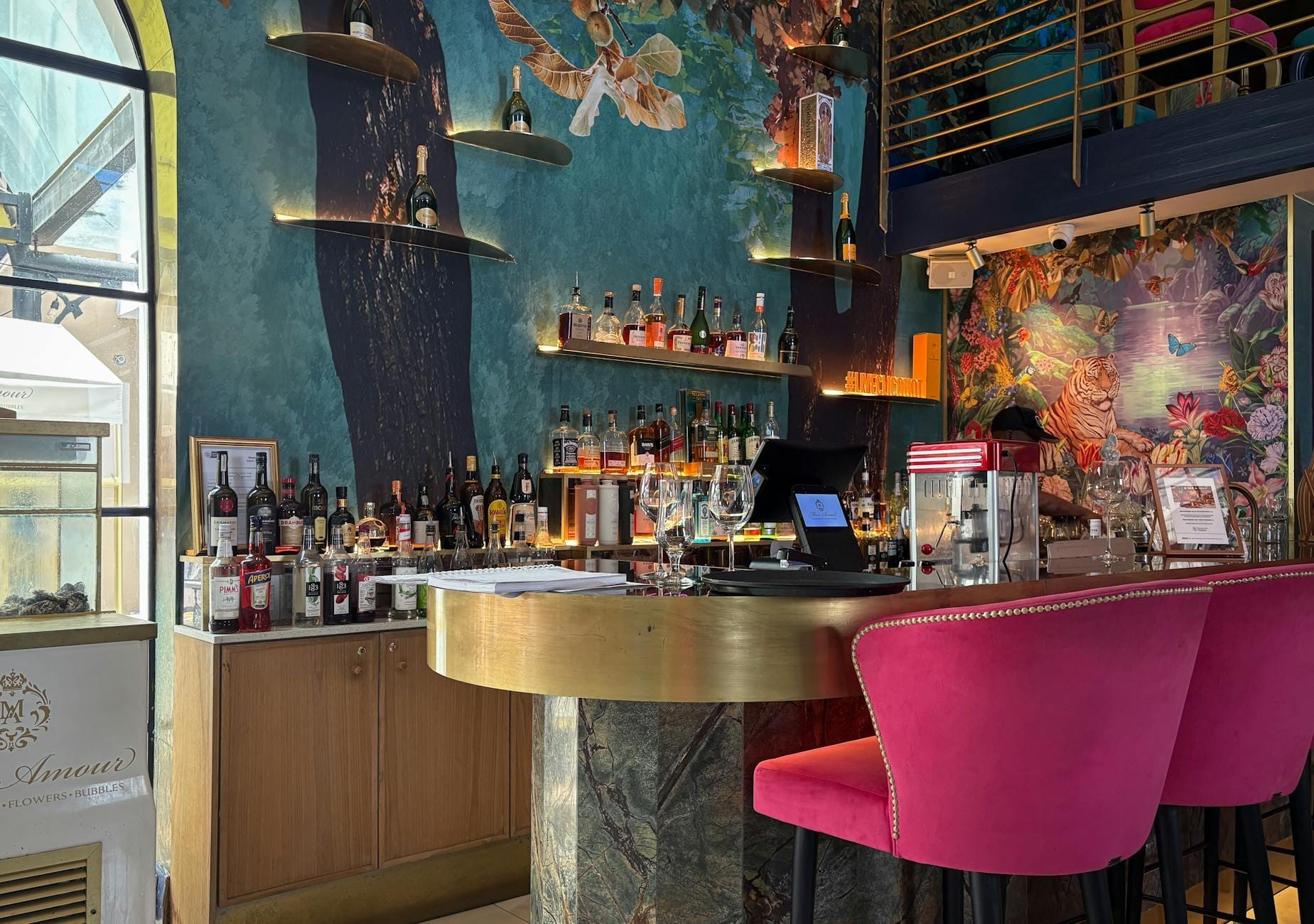 Bar de coquetéis chique e moderno em Nápoles com murais coloridos nas paredes, prateleiras de licores e confortáveis banquetas rosas em um ambiente animado e acolhedor.