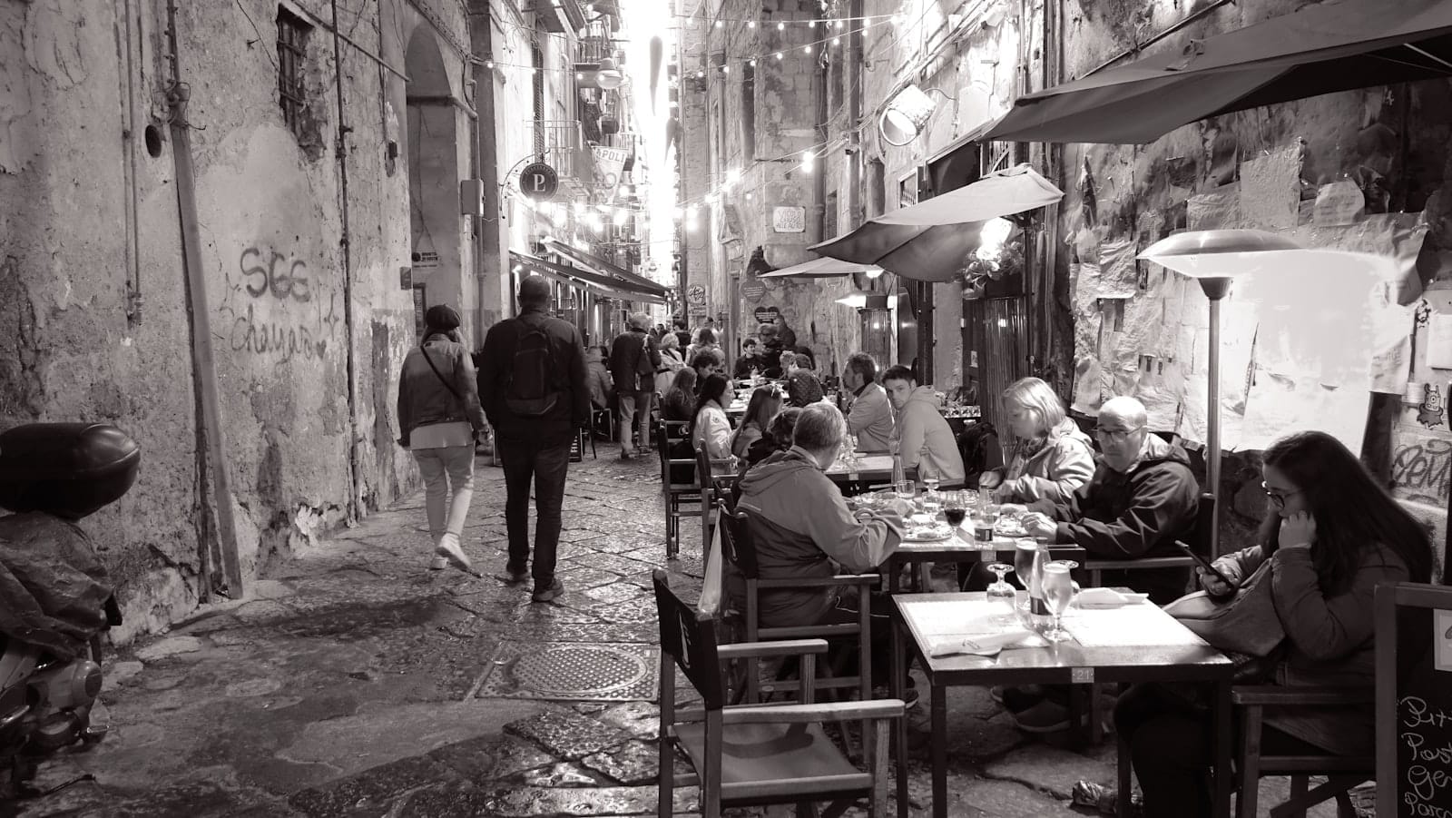 Cena noturna de rua em Nápoles com pessoas jantando ao ar livre em mesas de restaurante e cordas de luzes acima.