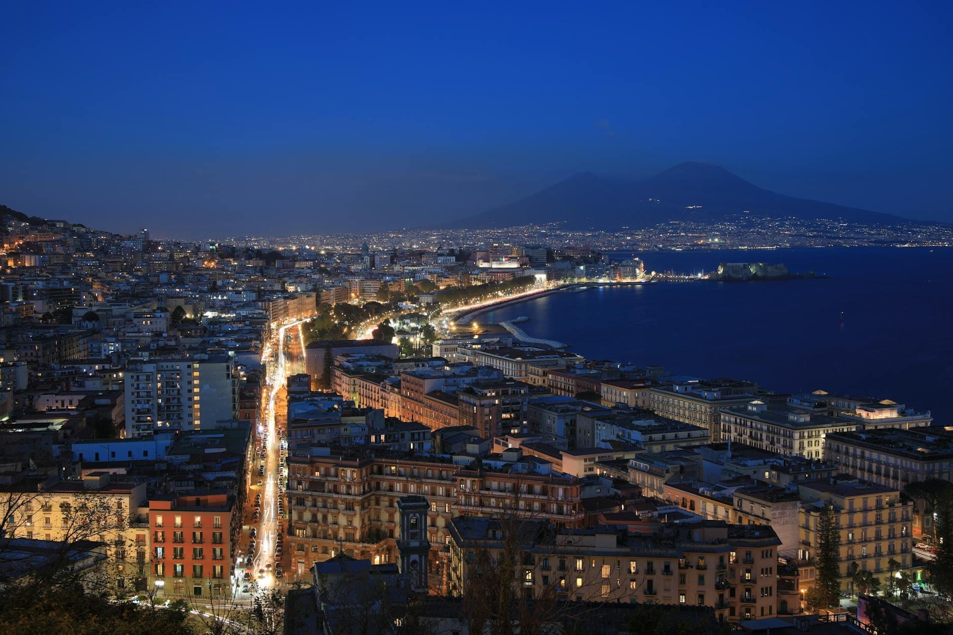 Vista panorâmica do litoral de Nápoles à noite com as luzes da cidade, ruas movimentadas e o Monte Vesúvio ao fundo sob um céu azul profundo.
