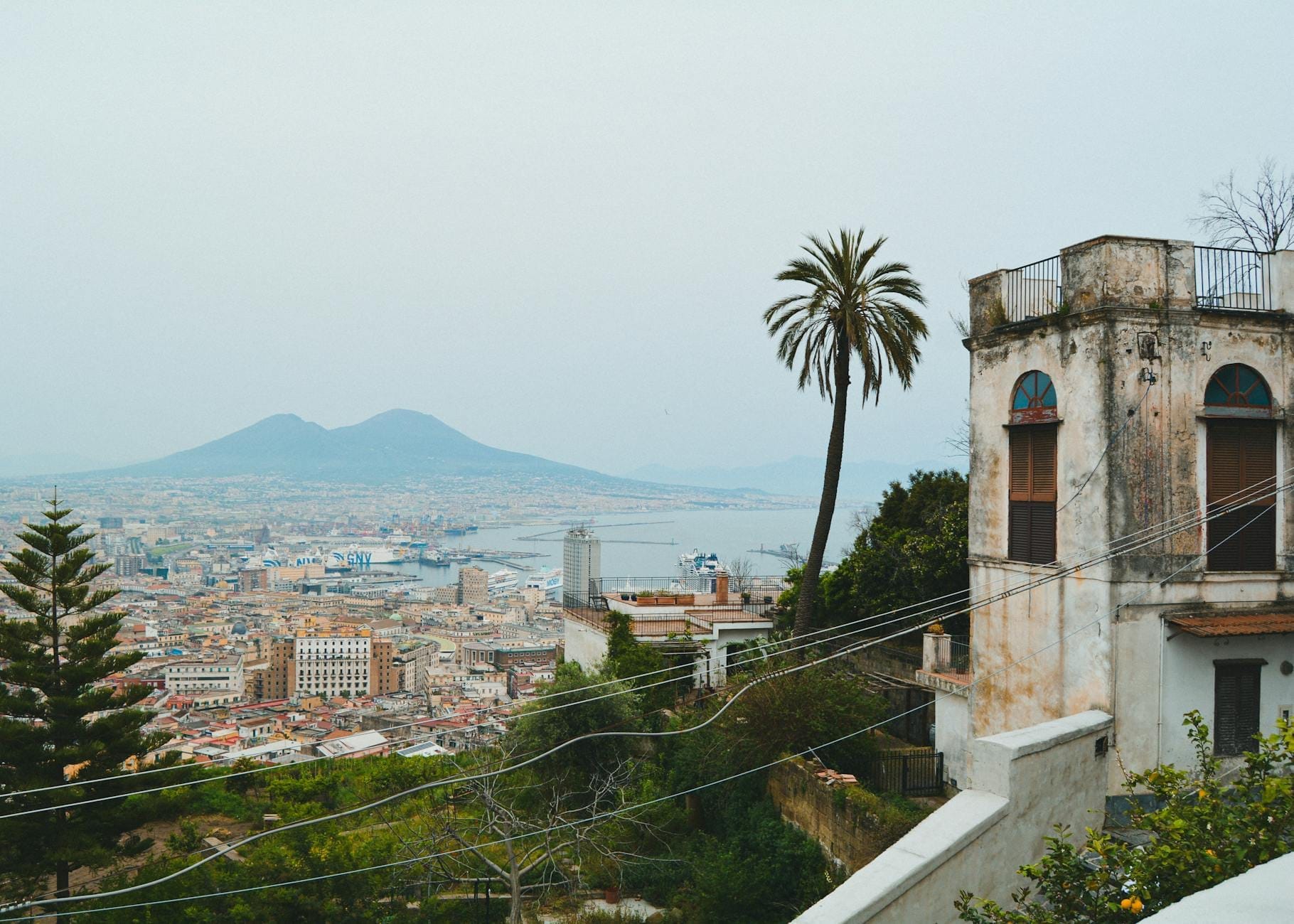 Vue sur Naples avec un vieux bâtiment, un palmier et le Mont Vésuve au loin, capturant l'atmosphère de la ville et la baie.