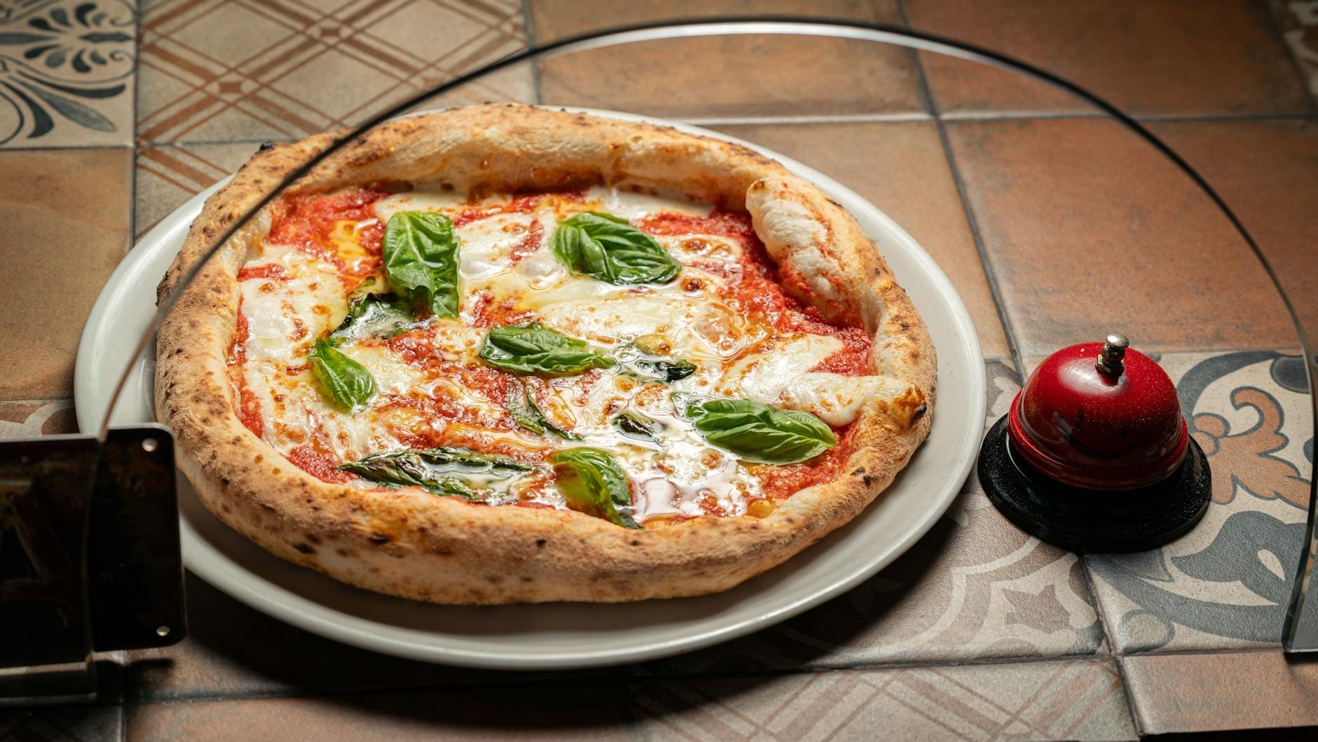 Pizza Margherita napolitaine classique dans une assiette avec du basilic frais, servie sur une table carrelée avec une cloche dans un cadre style pizzeria.