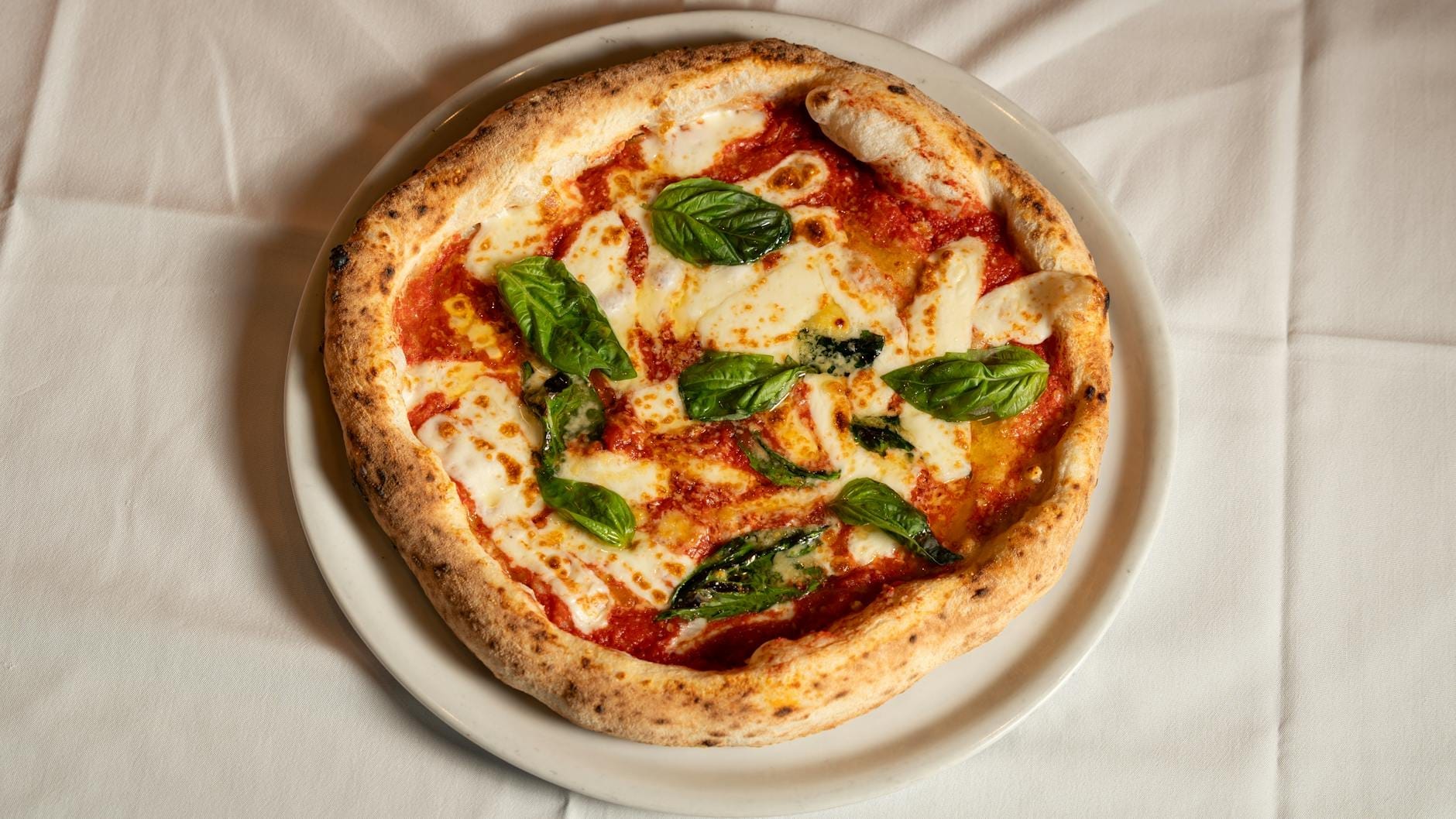 Pizza margherita napoletana clássica com manjericão fresco, mozzarella e molho de tomate, servida em prato branco sobre toalha branca, vista de cima.