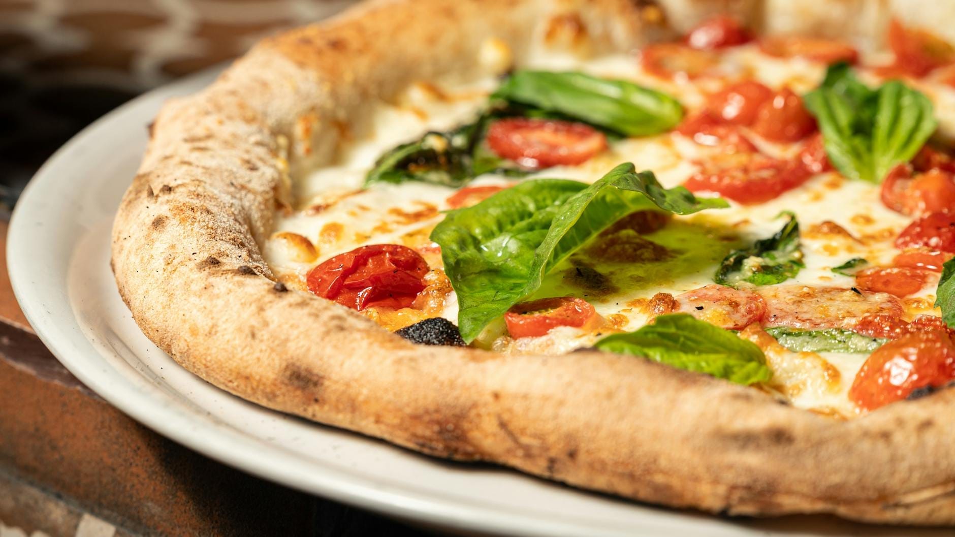 Primo piano di una classica pizza napoletana con basilico fresco, pomodorini e mozzarella su un piatto bianco, con uno sfondo di piastrelle rustiche.