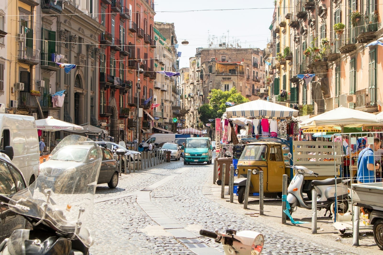 Scène de rue animée en journée à Naples avec des habitants, des voitures, des scooters, des étals de marché et des bâtiments traditionnels longeant une rue pavée.