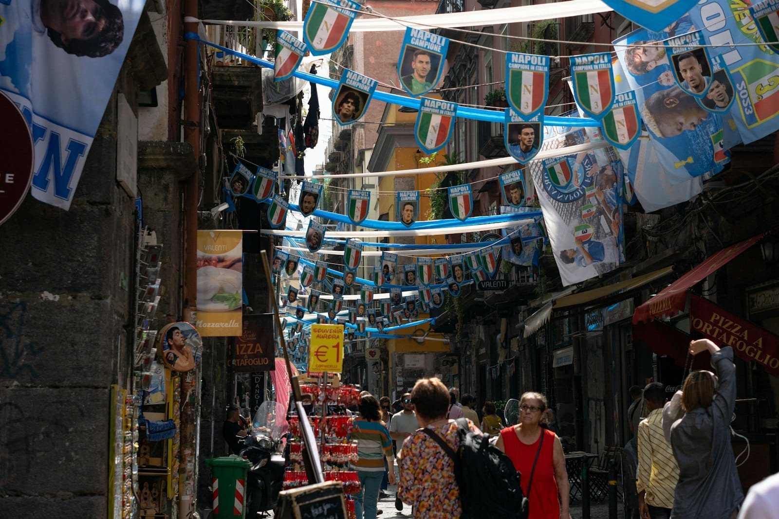 Une scène de rue animée à Naples avec des drapeaux et des banderoles, des personnes qui se promènent et des immeubles étroits serrés les uns contre les autres.