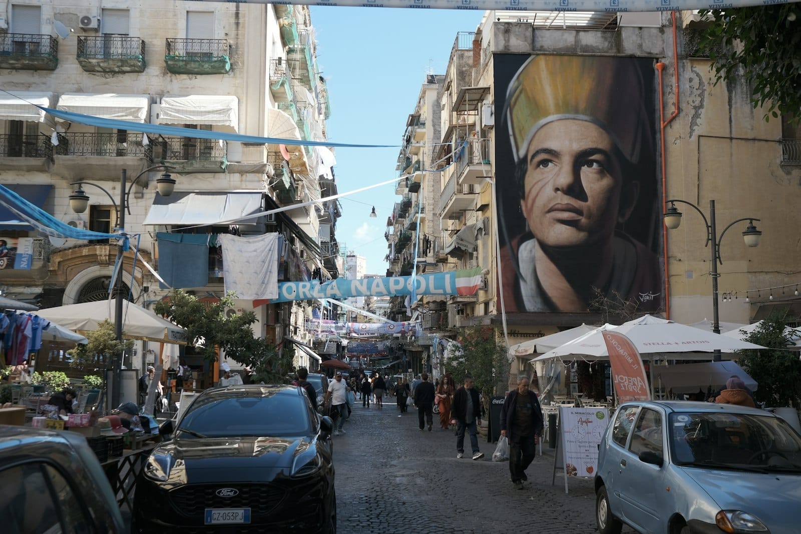 Movimentada rua de Nápoles com multidões, bancas ao ar livre e um mural de San Gennaro, mostrando uma celebração local perto do Duomo.