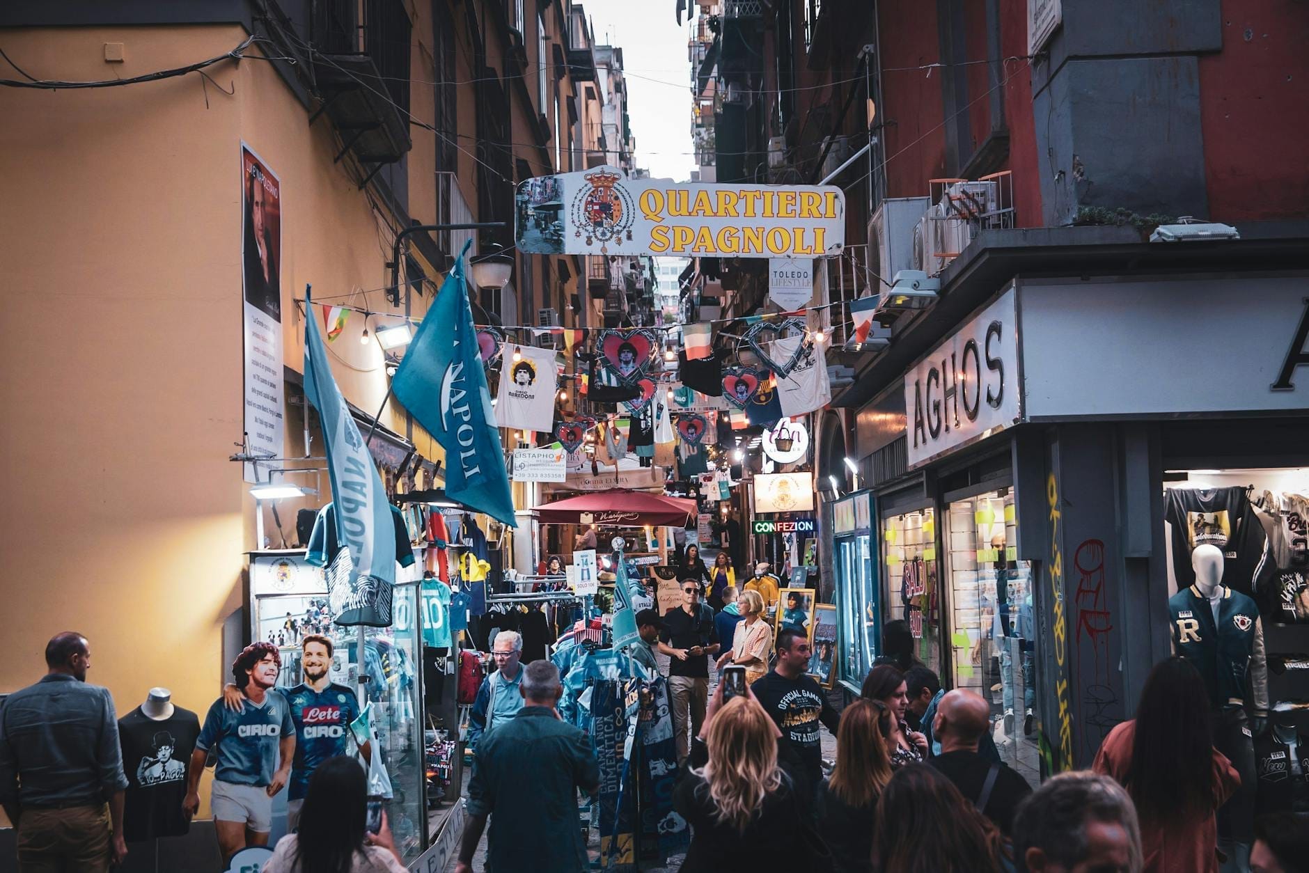 Uma movimentada rua estreita em Nápoles repleta de lojas, bancas de mercado, letreiros e multidões de pessoas, captando a atmosfera comercial local.