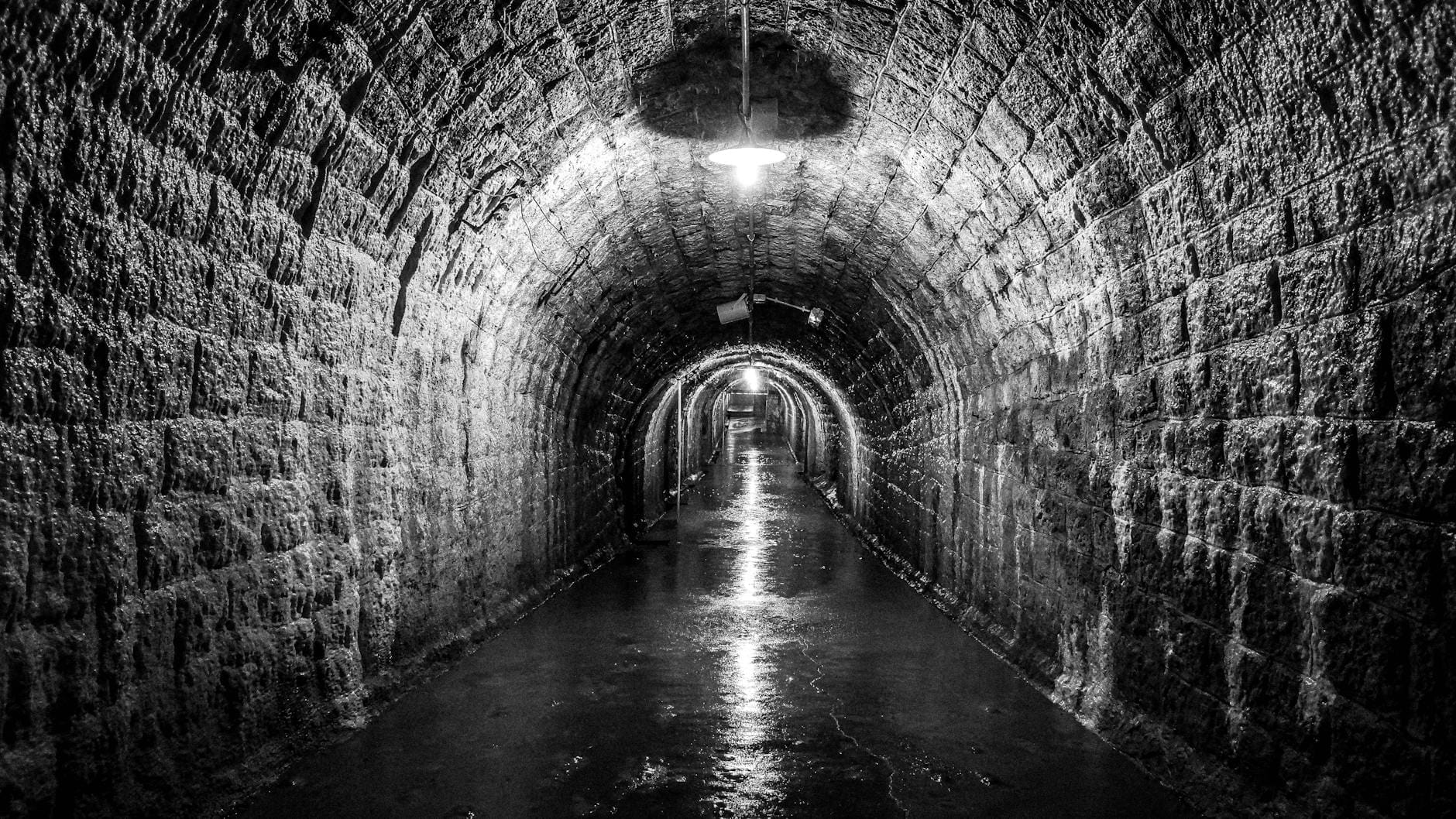 Photo en noir et blanc d'un tunnel en pierre avec un plafond voûté, un sol mouillé et une seule lumière suspendue au plafond.