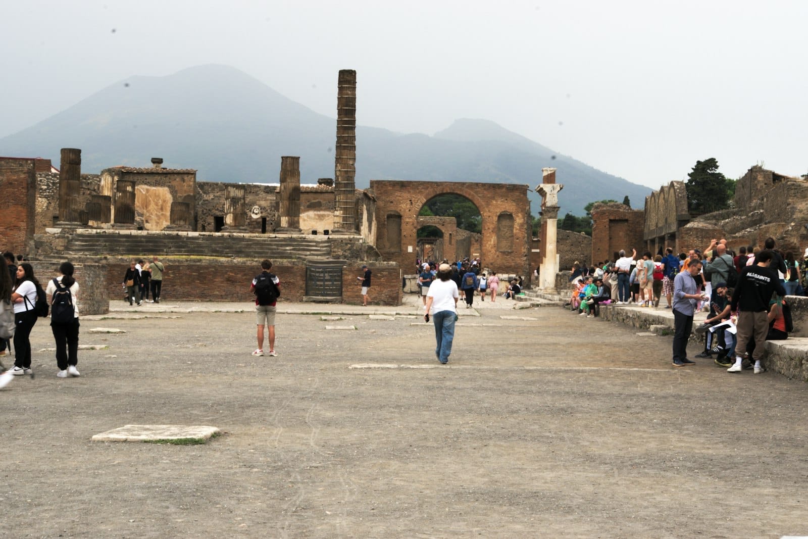 Des touristes se promènent parmi les ruines de Pompéi avec le Mont Vésuve visible en arrière-plan, zone du forum central.