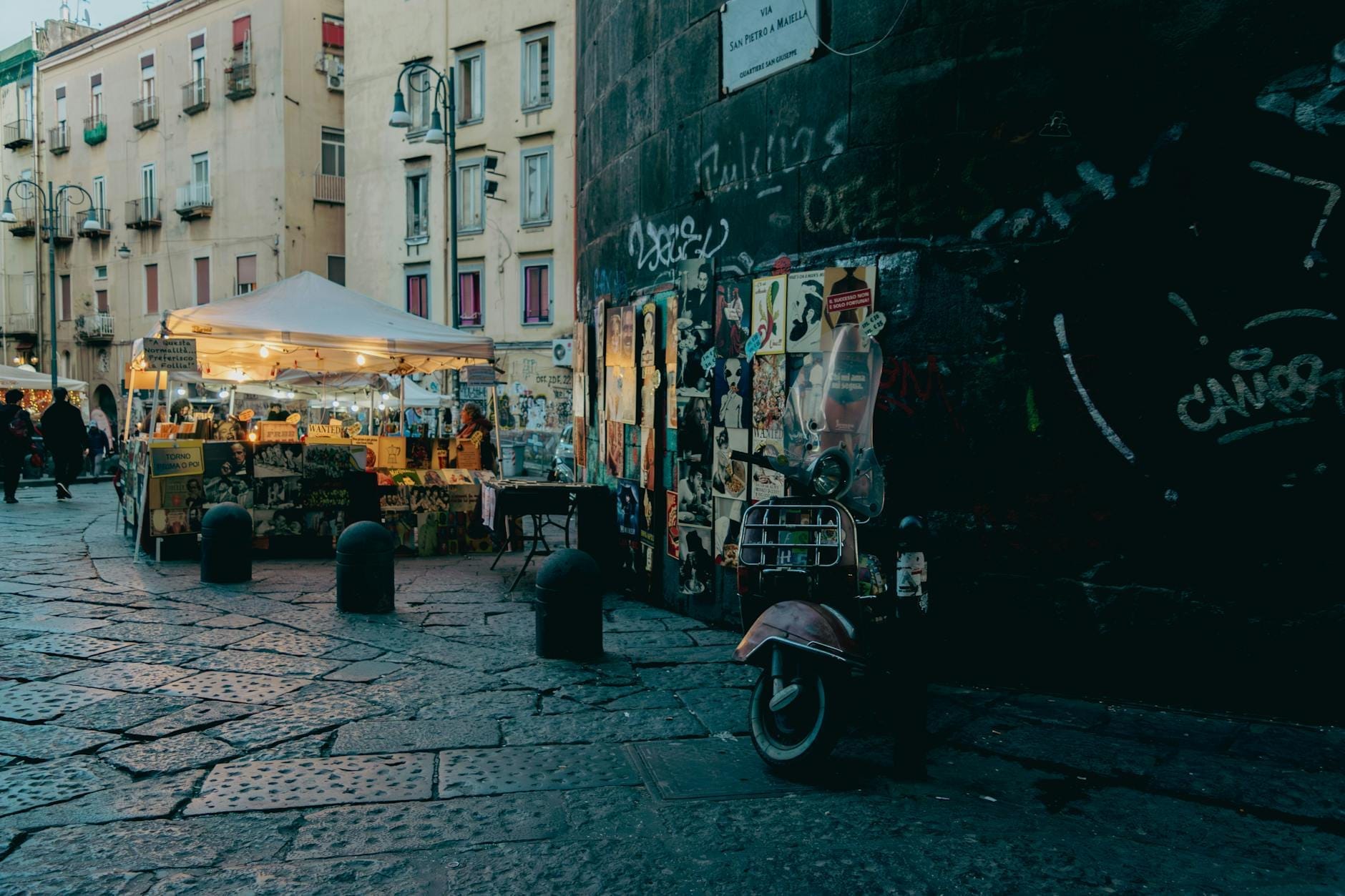 Scène de rue en soirée à Naples avec un stand de nourriture sous un auvent installé près de bâtiments urbains usés et un scooter garé.
