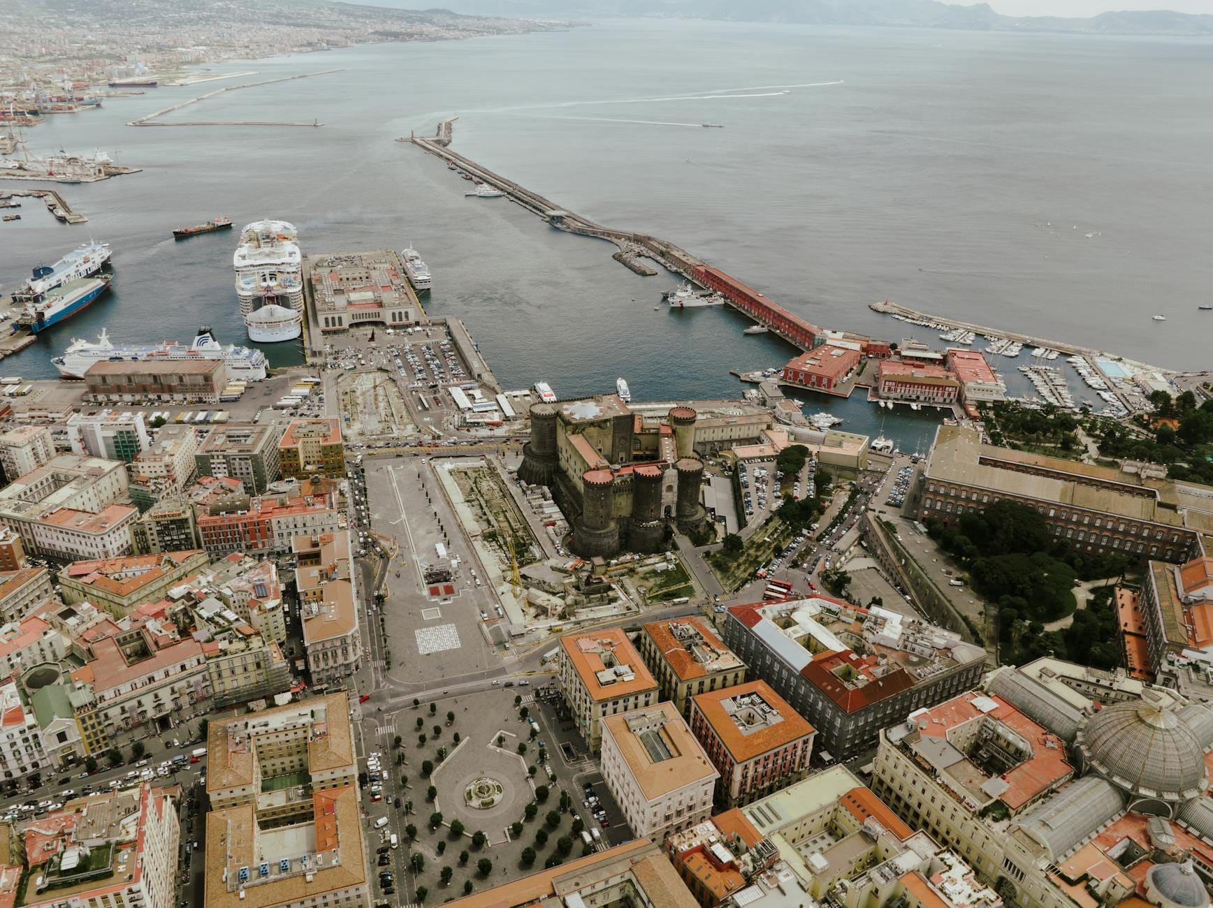Vue aérienne du port de Naples avec des ferries, des navires de croisière, des infrastructures portuaires et le Castello Maschio Angioino, montrant les principales options de transport de la ville.