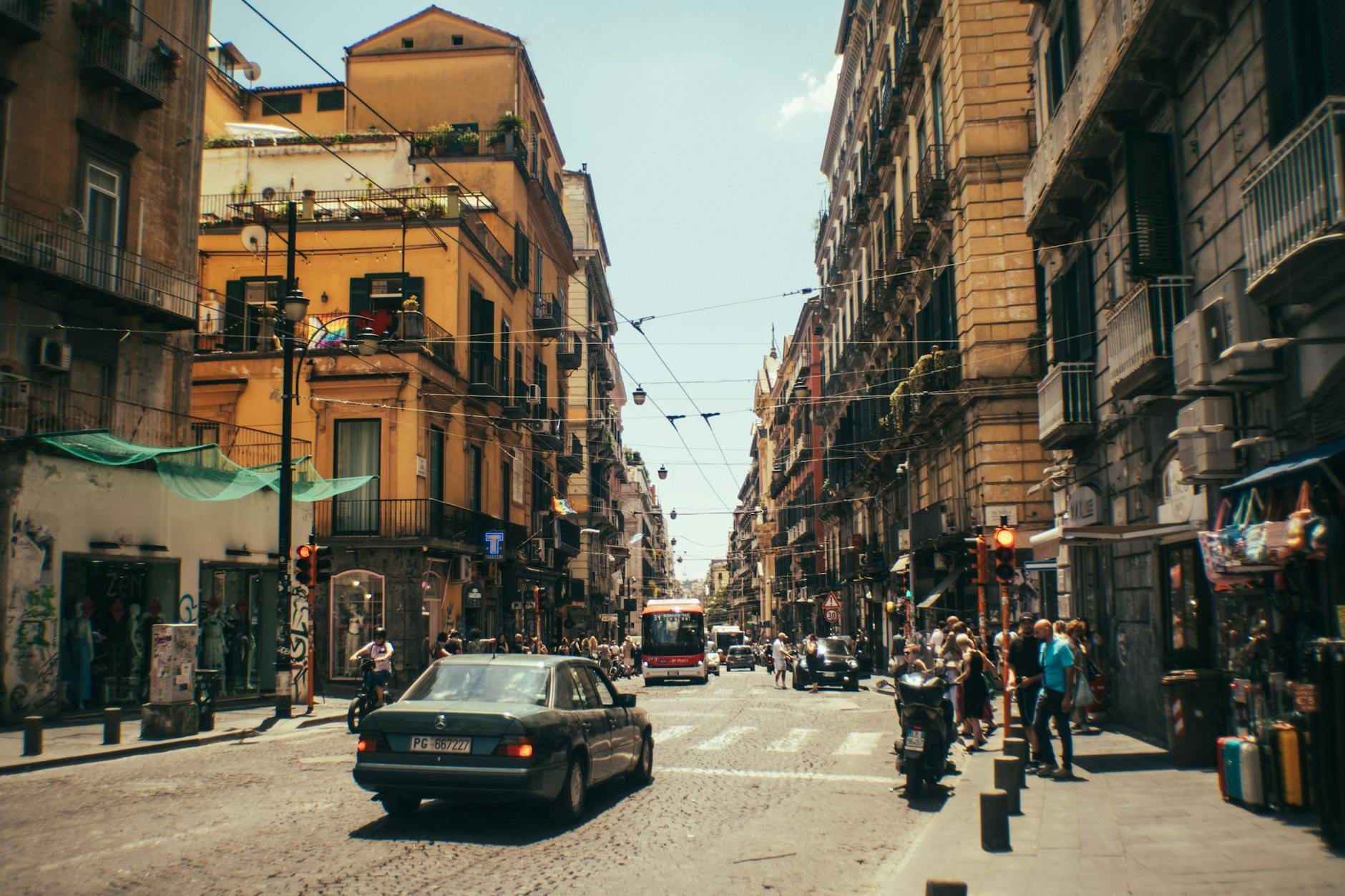 Rue animée de Naples avec voitures, passants et bâtiments ensoleillés, illustrant l'affluence urbaine et l'agitation saisonnière.