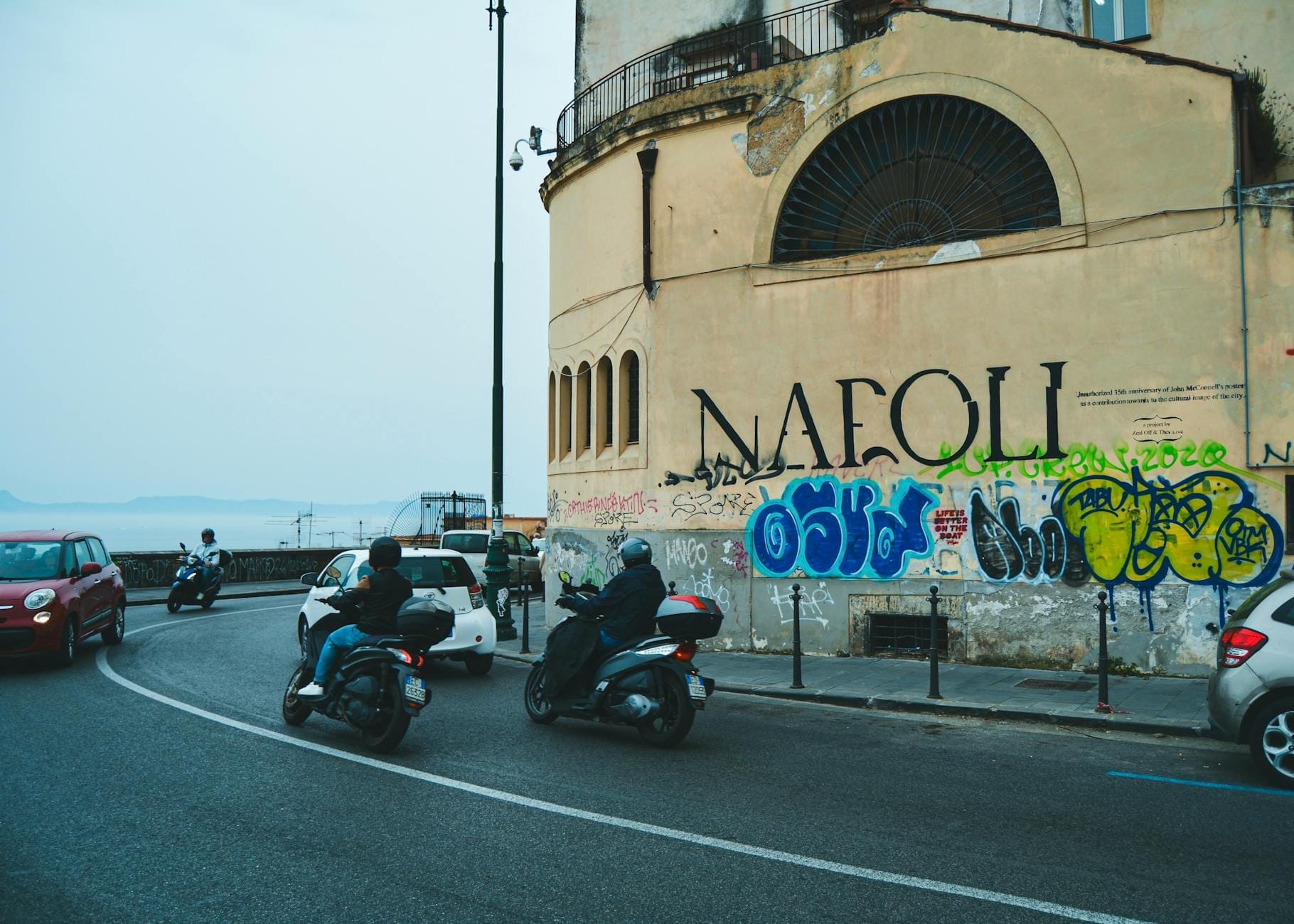 Scooters et voitures circulant dans une rue de Naples avec des graffitis, capturant la circulation locale et l'atmosphère urbaine.