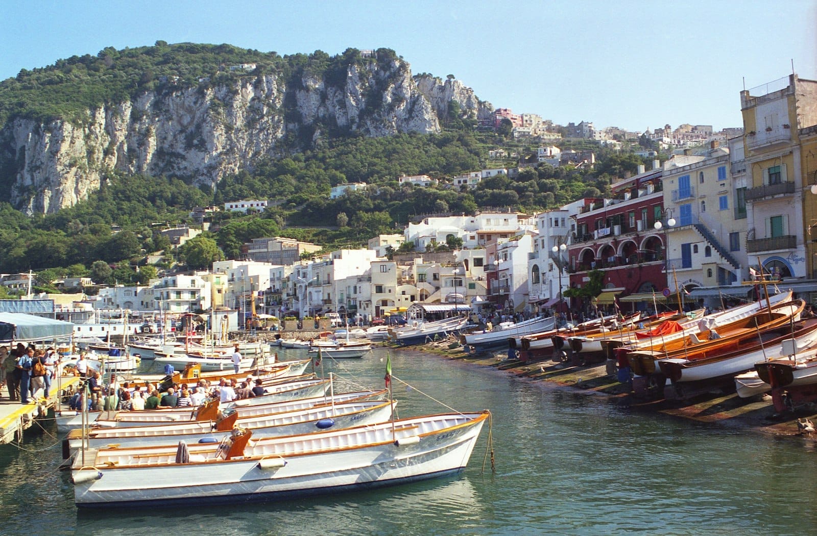 Barcos ancorados na charmosa marina de Capri com prédios coloridos e falésias dramáticas ao fundo em um dia ensolarado.