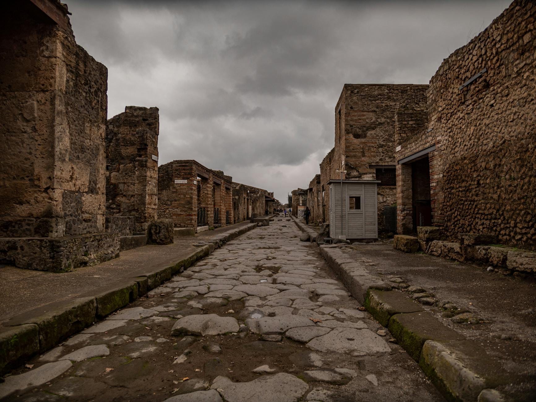 Antiga rua de pedra ladeada por ruínas de edifícios em Pompeia num dia nublado, mostrando a vastidão do sítio.