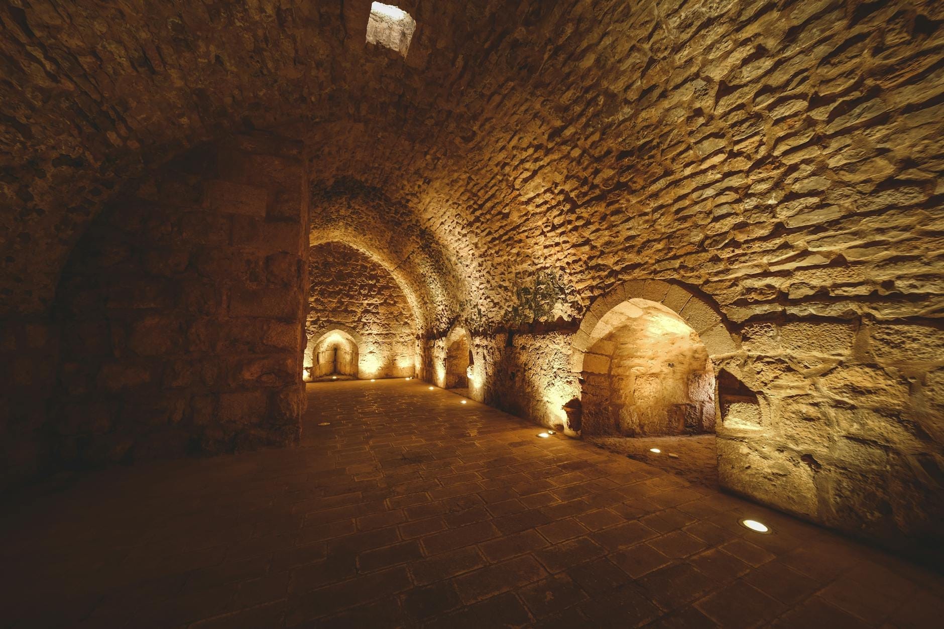 Passagem subterrânea em arco de pedra com nichos iluminados, mostrando a estrutura de uma antiga câmara subterrânea.